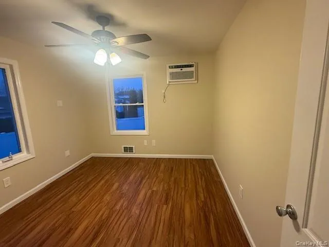 Empty room with dark wood-style flooring, ceiling fan, and an AC wall unit Empty room with dark wood-style flooring, ceiling fan, and an AC wall unit