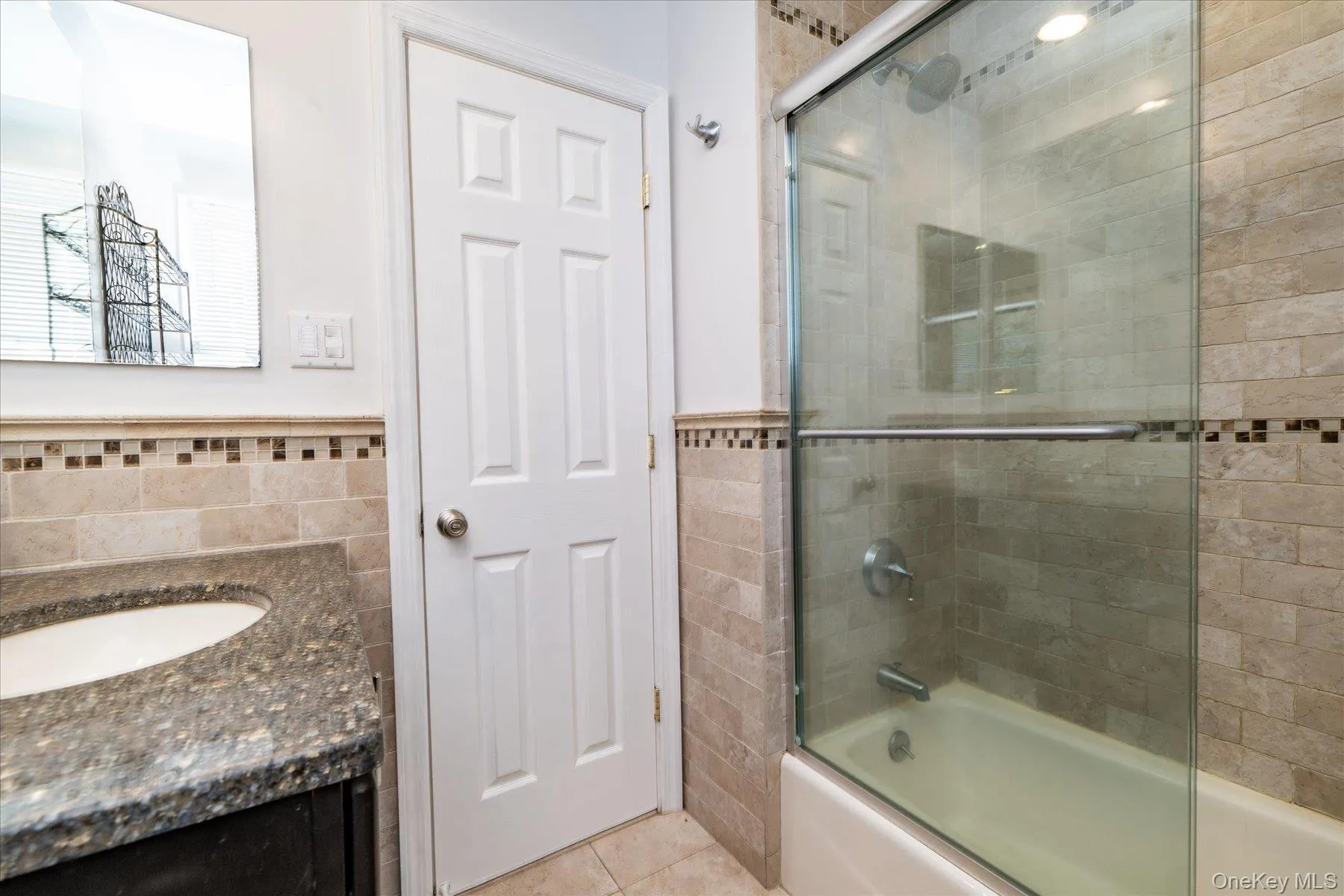 Full bath with vanity, shower / bath combination with glass door, tile walls, light tile patterned floors, and wainscoting Full bath with vanity, shower / bath combination with glass door, tile walls, light tile patterned floors, and wainscoting