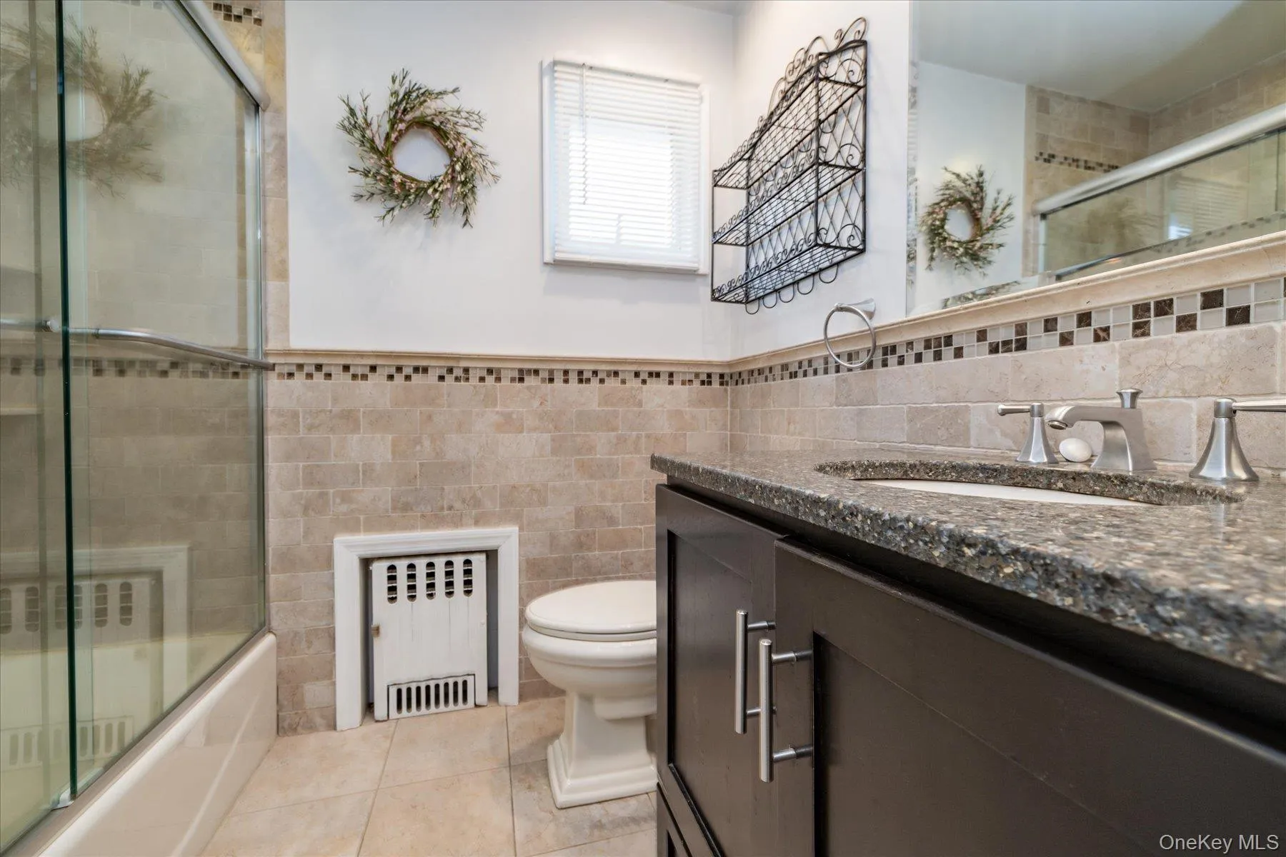 Full bathroom with vanity, radiator, tile walls, light tile patterned flooring, and bath / shower combo with glass door Full bathroom with vanity, radiator, tile walls, light tile patterned flooring, and bath / shower combo with glass door