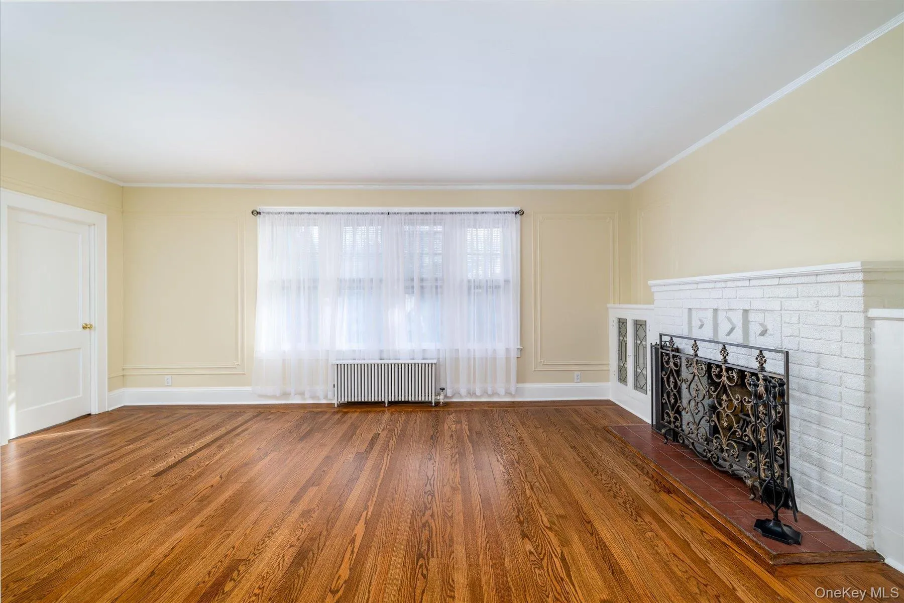 Unfurnished living room with hardwood / wood-style floors, radiator heating unit, a brick fireplace, a decorative wall, and ornamental molding Unfurnished living room with hardwood / wood-style floors, radiator heating unit, a brick fireplace, a decorative wall, and ornamental molding