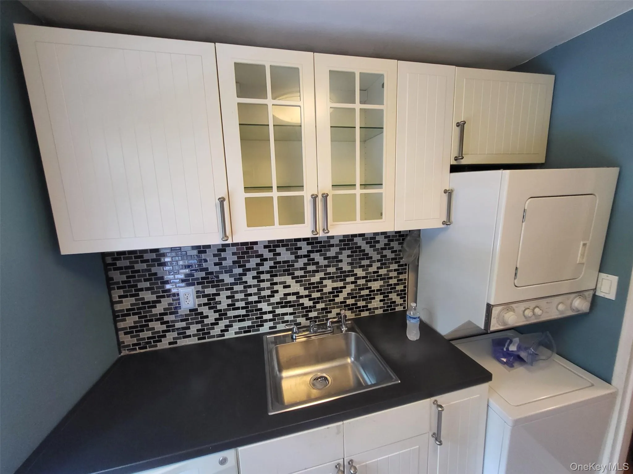 Kitchen with dark countertops, stacked washing machine and dryer, tasteful backsplash, and white cabinetry Kitchen with dark countertops, stacked washing machine and dryer, tasteful backsplash, and white cabinetry