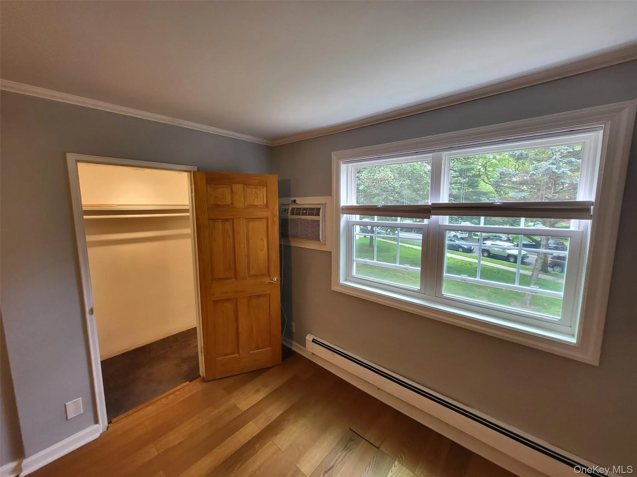 Unfurnished bedroom featuring baseboard heating, light wood-type flooring, ornamental molding, a wall mounted AC, and a closet Unfurnished bedroom featuring baseboard heating, light wood-type flooring, ornamental molding, a wall mounted AC, and a closet