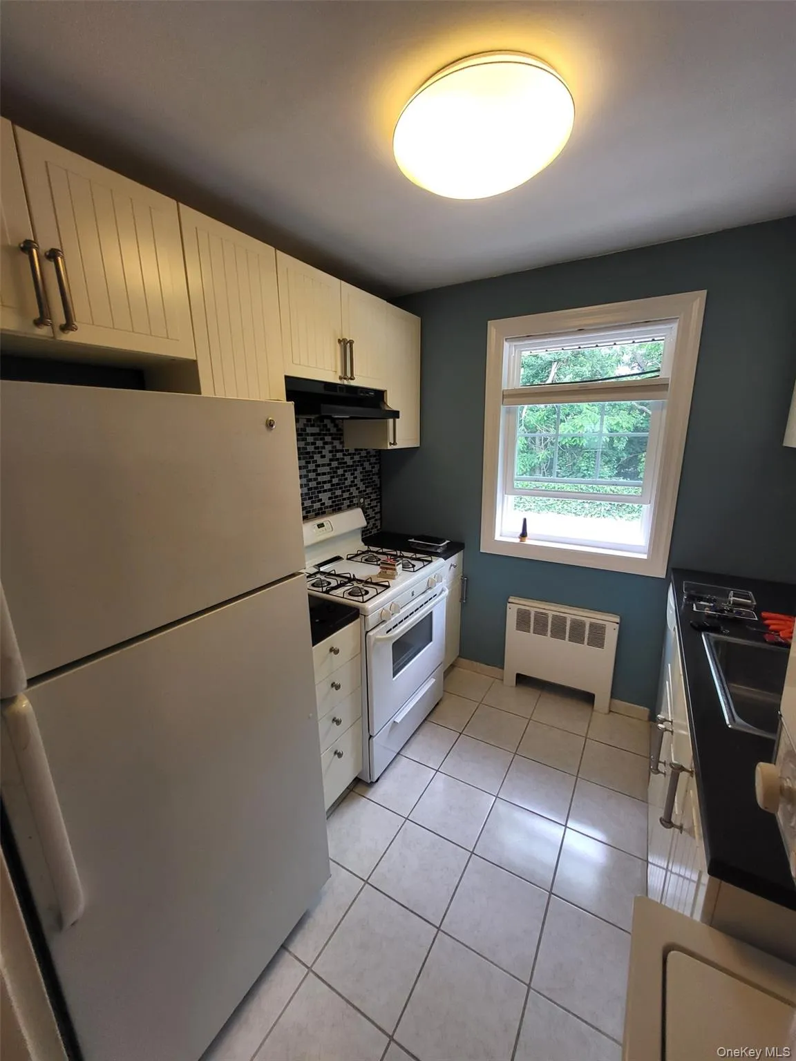 Kitchen with white appliances, radiator heating unit, under cabinet range hood, tasteful backsplash, and light tile patterned flooring Kitchen with white appliances, radiator heating unit, under cabinet range hood, tasteful backsplash, and light tile patterned flooring