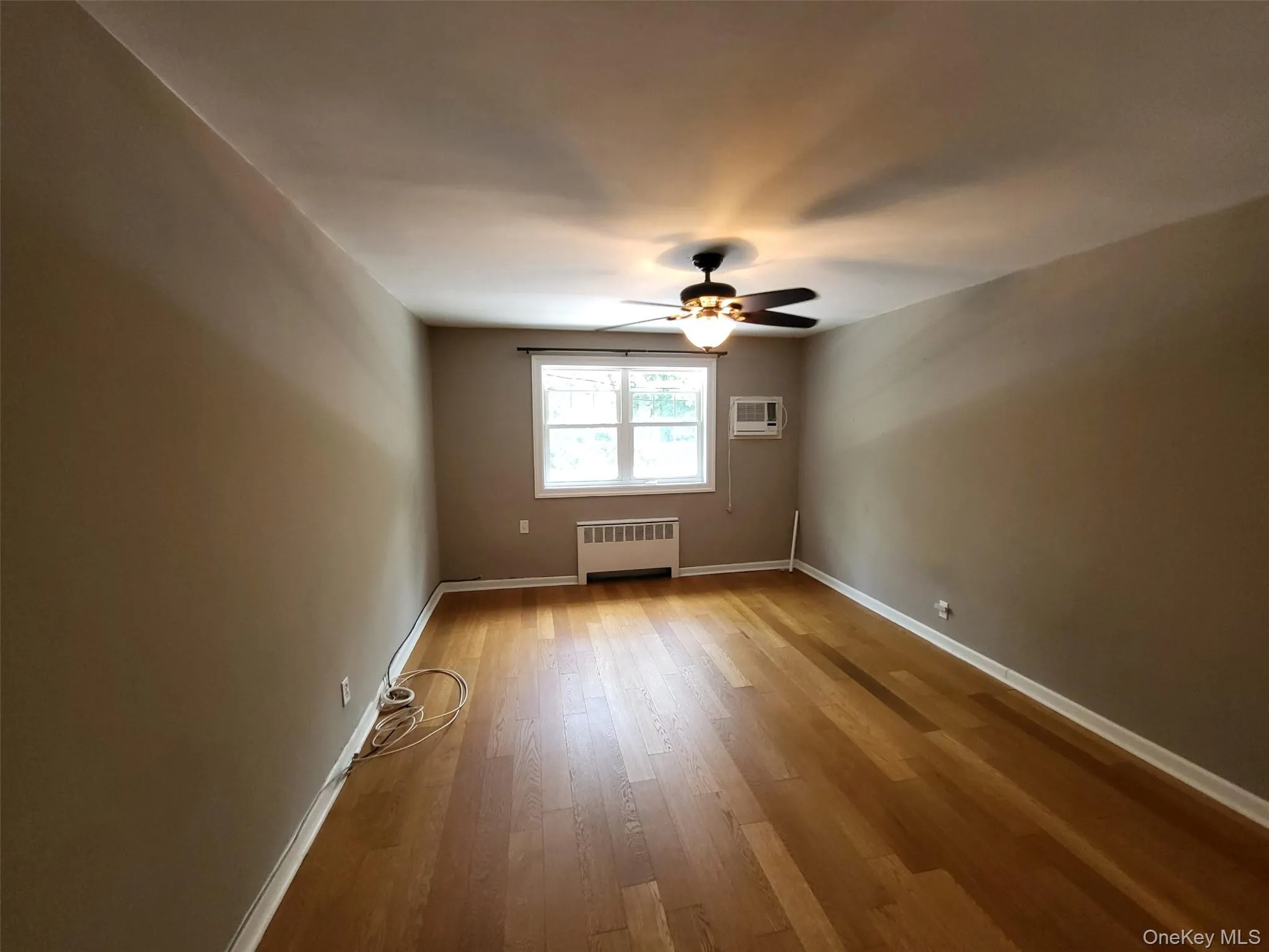 Spare room with radiator, an AC wall unit, ceiling fan, and wood finished floors Spare room with radiator, an AC wall unit, ceiling fan, and wood finished floors