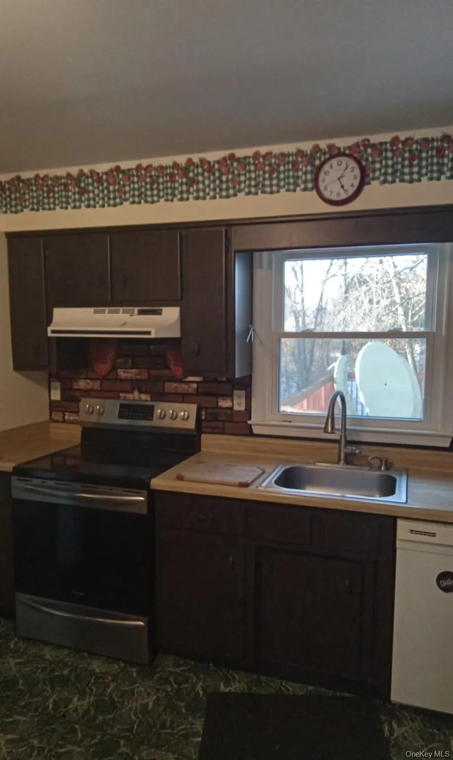 Kitchen with dark brown cabinets, electric stove, under cabinet range hood, white dishwasher, and light countertops Kitchen with dark brown cabinets, electric stove, under cabinet range hood, white dishwasher, and light countertops