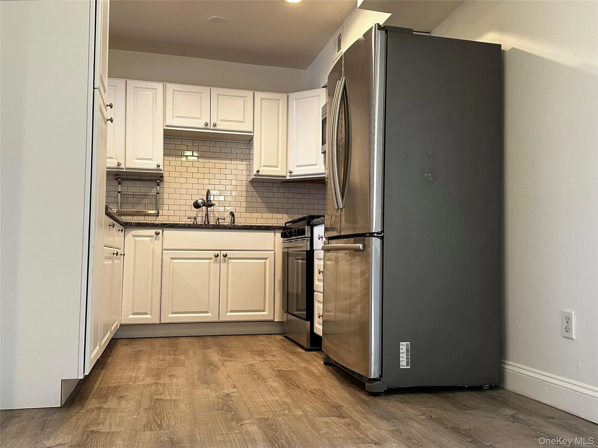 Kitchen with appliances with stainless steel finishes, white cabinetry, tasteful backsplash, dark stone countertops, and vinyl floors Kitchen with appliances with stainless steel finishes, white cabinetry, tasteful backsplash, dark stone countertops, and vinyl floors