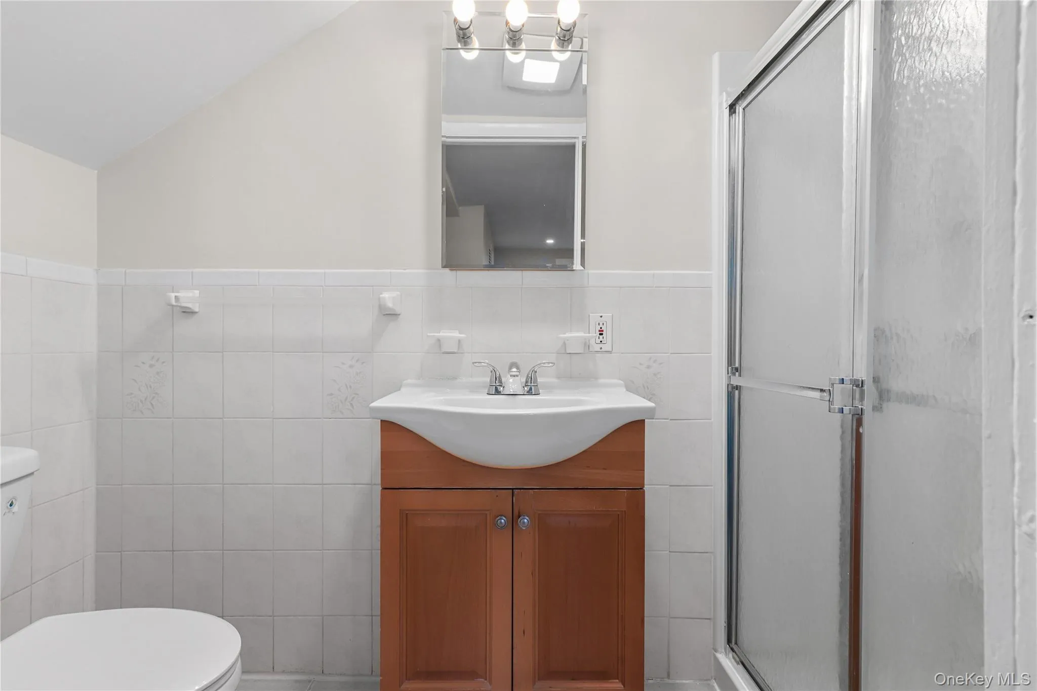 Bathroom featuring a stall shower, vanity, tile walls, a wainscoted wall, and lofted ceiling Bathroom featuring a stall shower, vanity, tile walls, a wainscoted wall, and lofted ceiling