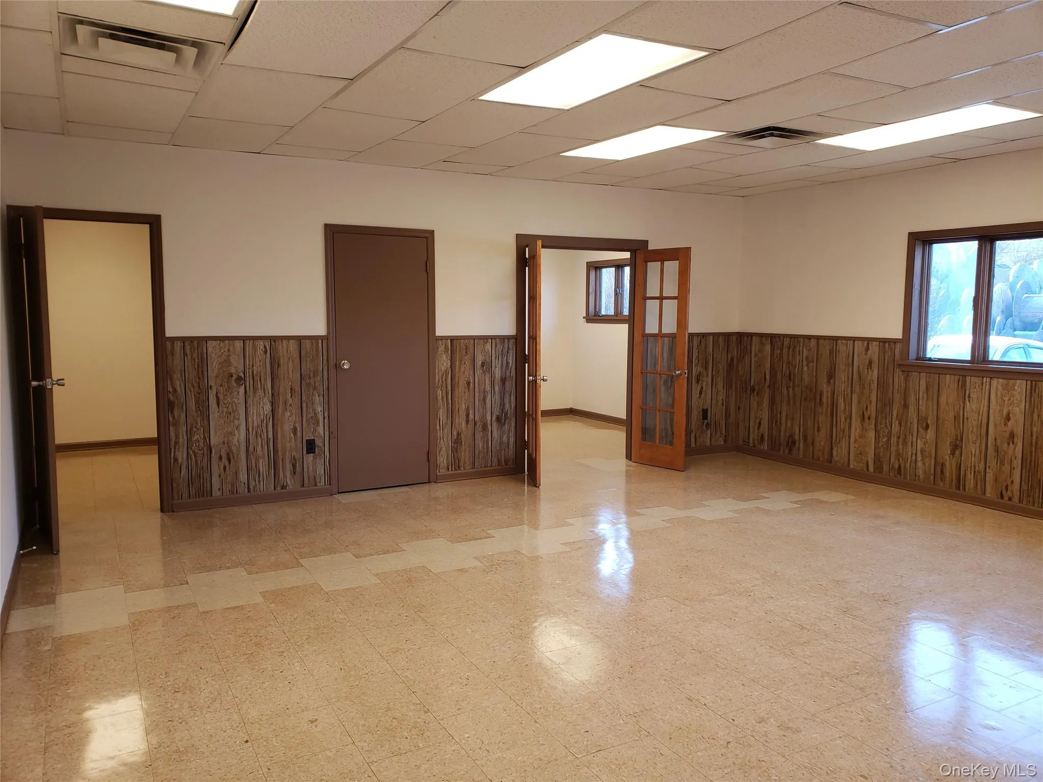 Empty room featuring light flooring, wood walls, french doors, wainscoting, and a drop ceiling Empty room featuring light flooring, wood walls, french doors, wainscoting, and a drop ceiling