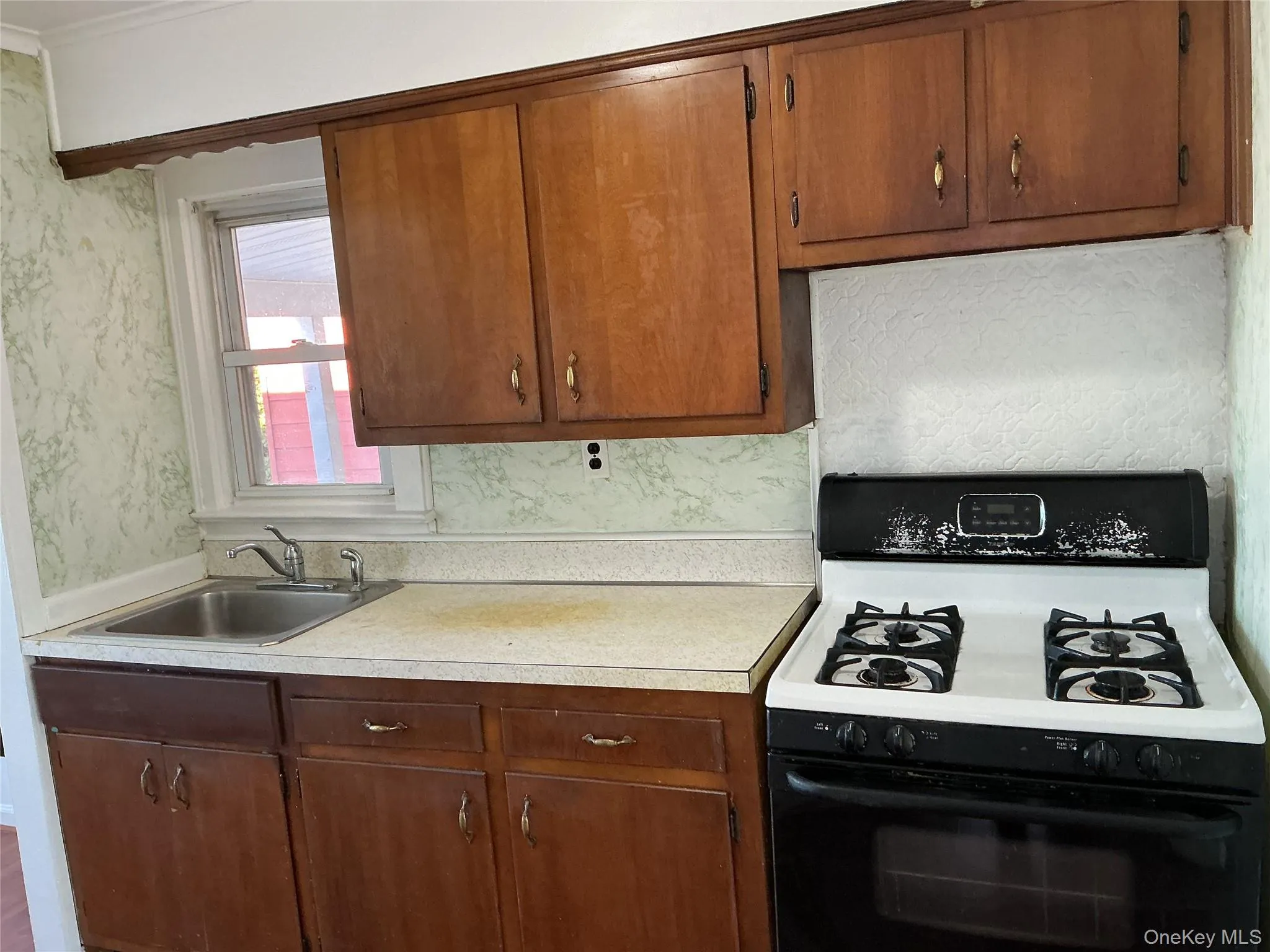 Kitchen featuring range with gas stovetop, light countertops, wallpapered walls, and brown cabinetry Kitchen featuring range with gas stovetop, light countertops, wallpapered walls, and brown cabinetry