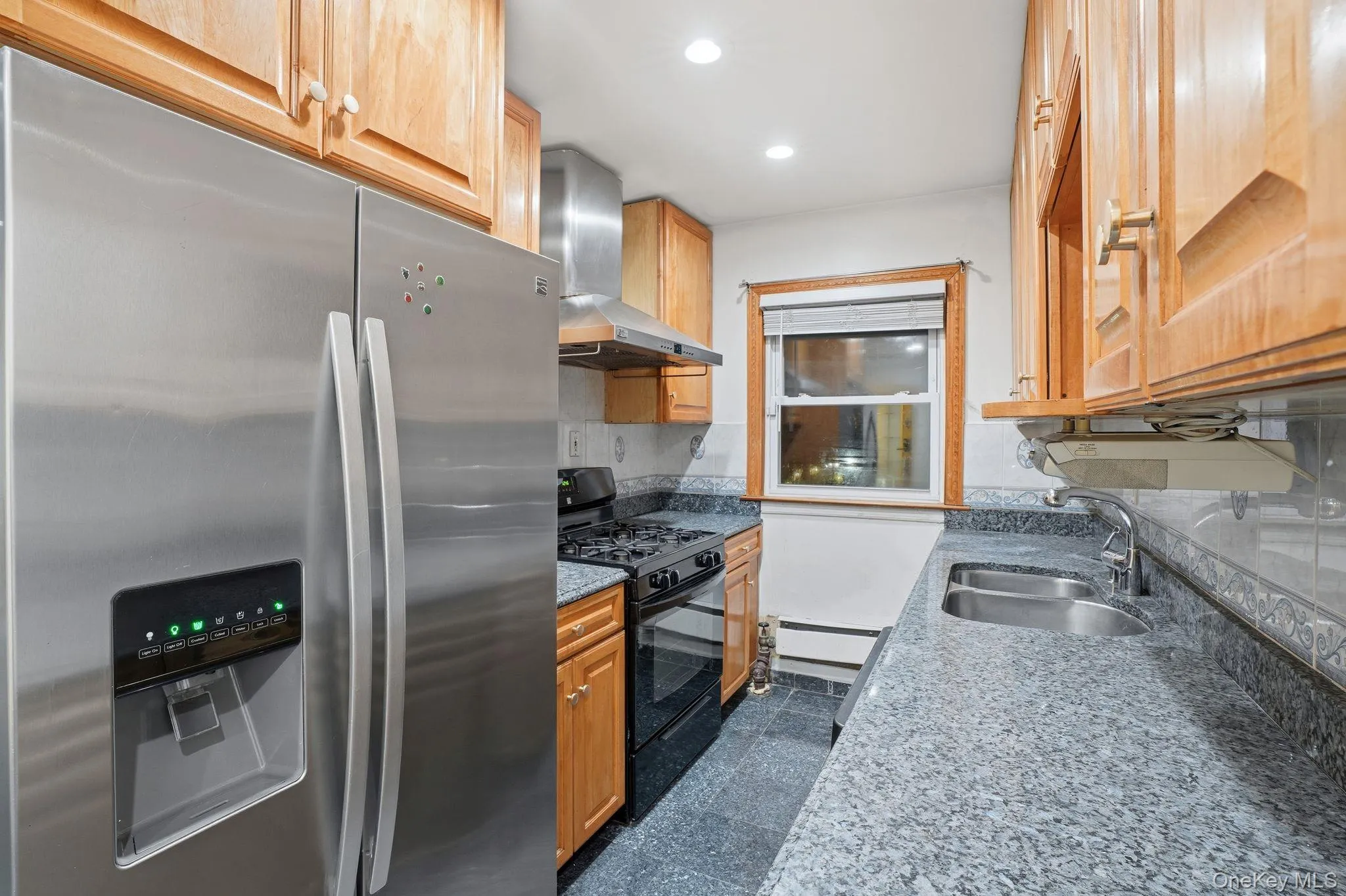 Kitchen with stainless steel refrigerator with ice dispenser, black range with gas cooktop, dark stone counters, wall chimney exhaust hood, and recessed lighting Kitchen with stainless steel refrigerator with ice dispenser, black range with gas cooktop, dark stone counters, wall chimney exhaust hood, and recessed lighting