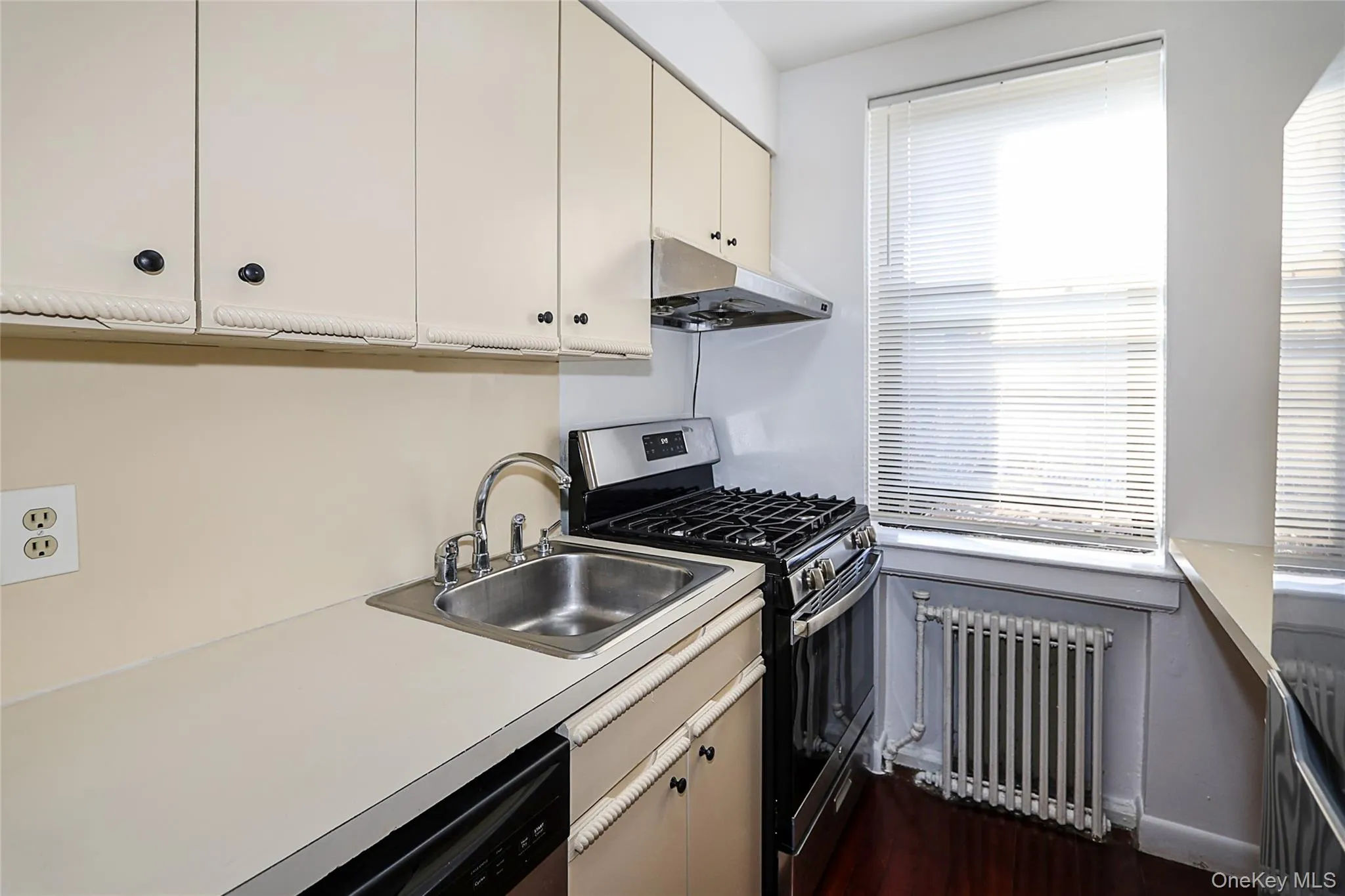Kitchen with stainless steel appliances, radiator heating unit, light countertops, under cabinet range hood, and dark wood-style floors Kitchen with stainless steel appliances, radiator heating unit, light countertops, under cabinet range hood, and dark wood-style floors