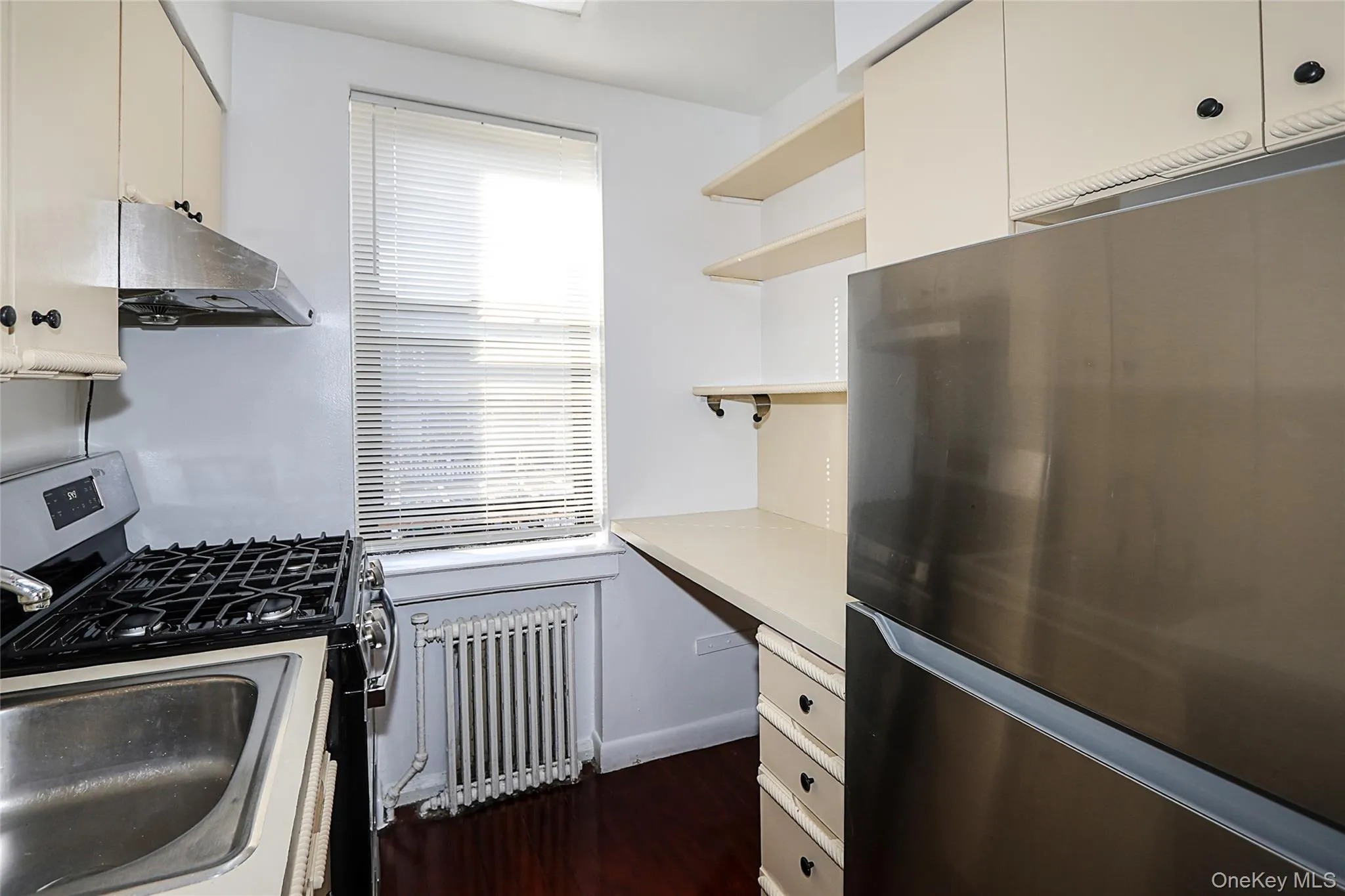 Kitchen featuring appliances with stainless steel finishes, open shelves, radiator heating unit, light countertops, and under cabinet range hood Kitchen featuring appliances with stainless steel finishes, open shelves, radiator heating unit, light countertops, and under cabinet range hood