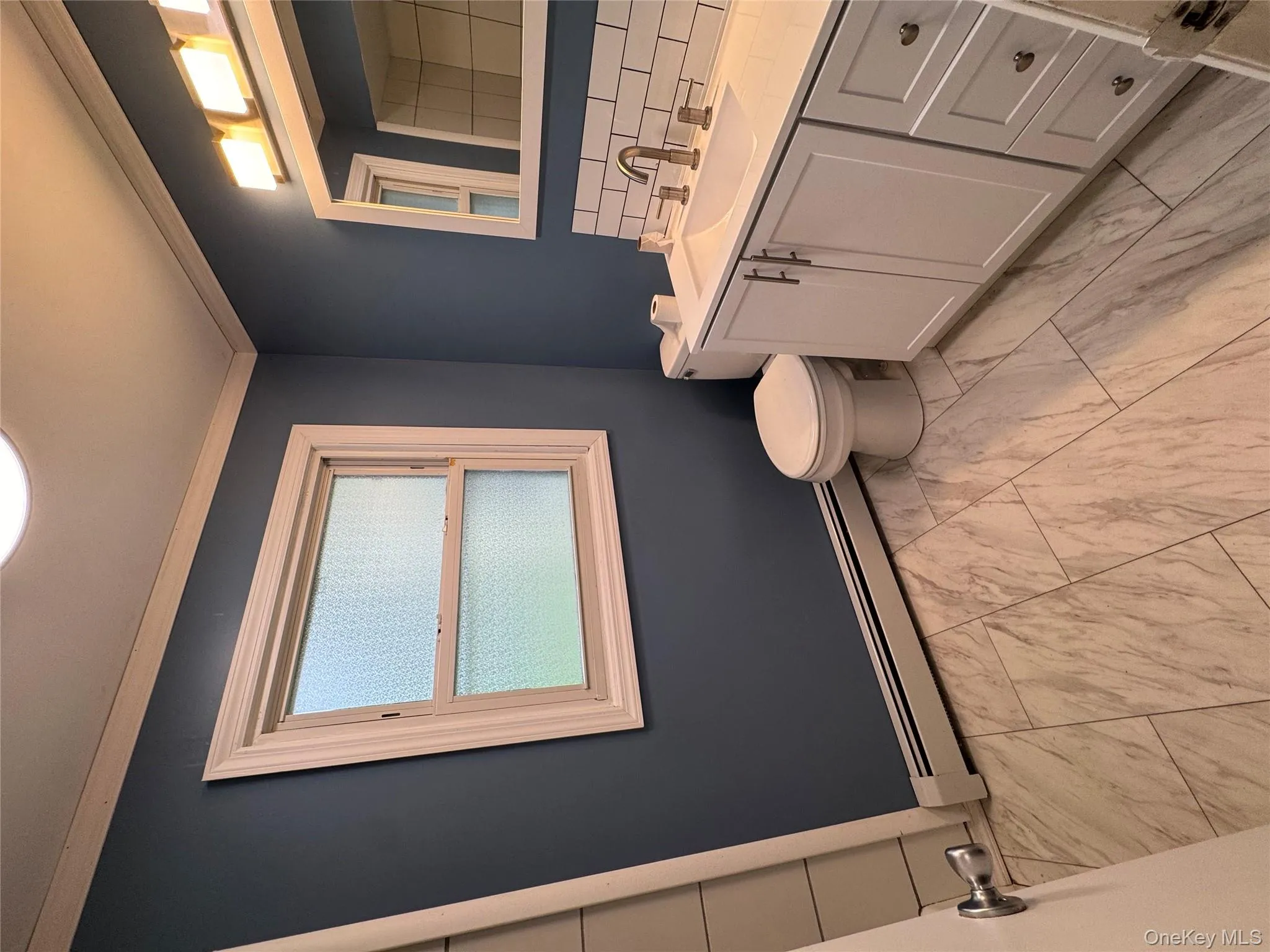 Bathroom featuring vanity, a baseboard radiator, decorative backsplash, ornamental molding, and light marble finish floors Bathroom featuring vanity, a baseboard radiator, decorative backsplash, ornamental molding, and light marble finish floors