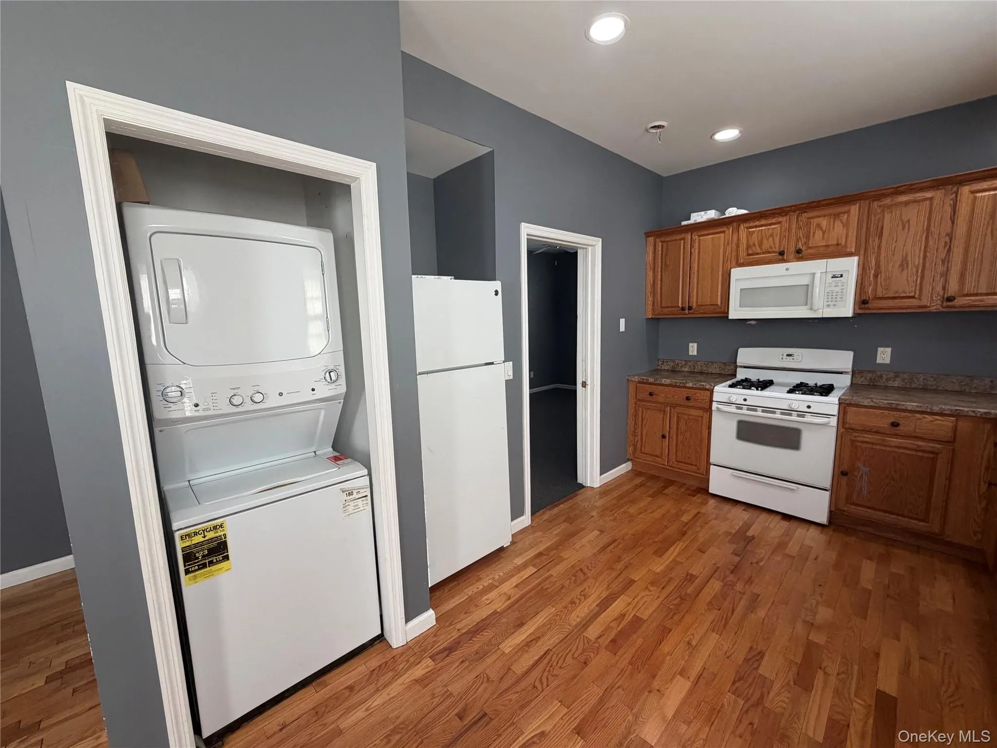Kitchen with white appliances, light wood-style floors, stacked washer / drying machine, dark countertops, and recessed lighting Kitchen with white appliances, light wood-style floors, stacked washer / drying machine, dark countertops, and recessed lighting