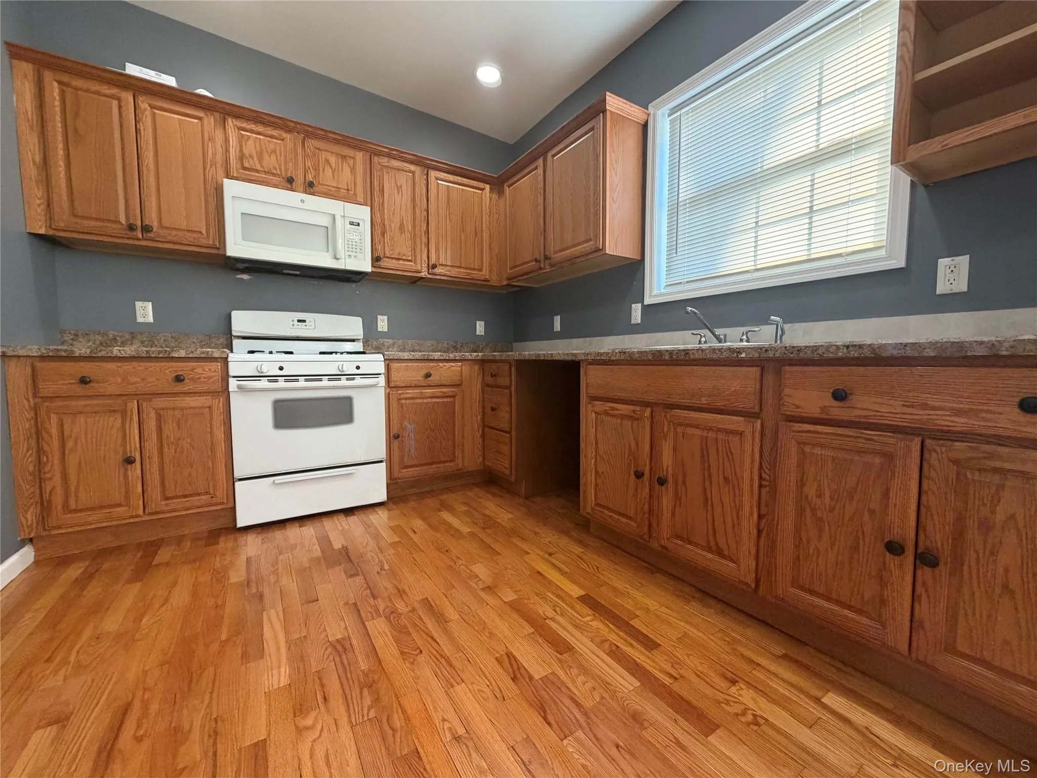Kitchen with white appliances, brown cabinetry, light wood finished floors, recessed lighting, and dark stone counters Kitchen with white appliances, brown cabinetry, light wood finished floors, recessed lighting, and dark stone counters