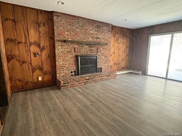 Unfurnished living room with wood walls, a fireplace, wood finished floors, and a baseboard radiator Unfurnished living room with wood walls, a fireplace, wood finished floors, and a baseboard radiator