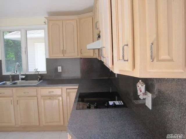 Kitchen with light brown cabinetry, black electric stovetop, backsplash, range hood, and dark countertops Kitchen with light brown cabinetry, black electric stovetop, backsplash, range hood, and dark countertops