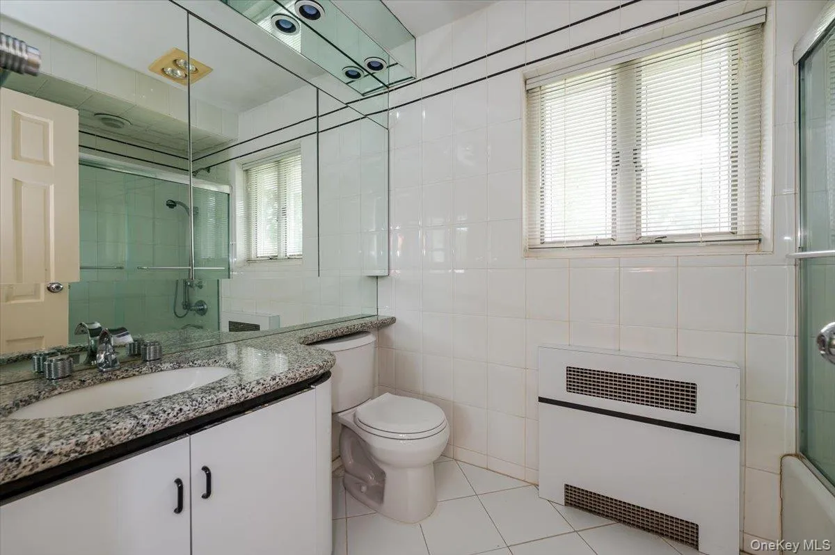 Bathroom featuring vanity, light tile patterned floors, heating unit, shower / bath combination with glass door, and tile walls Bathroom featuring vanity, light tile patterned floors, heating unit, shower / bath combination with glass door, and tile walls