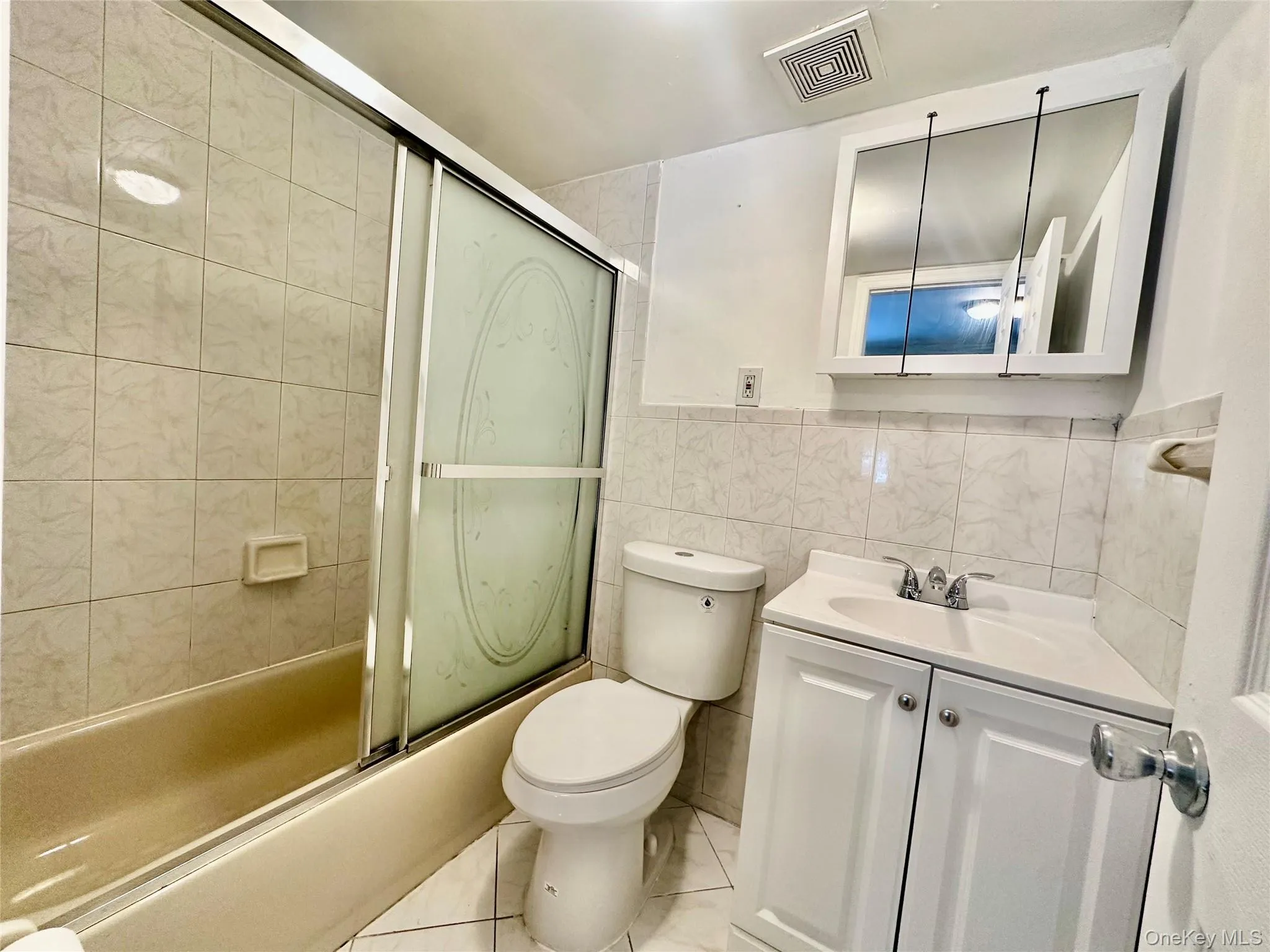 Full bathroom with tile walls, vanity, combined bath / shower with glass door, light tile patterned flooring, and wainscoting Full bathroom with tile walls, vanity, combined bath / shower with glass door, light tile patterned flooring, and wainscoting