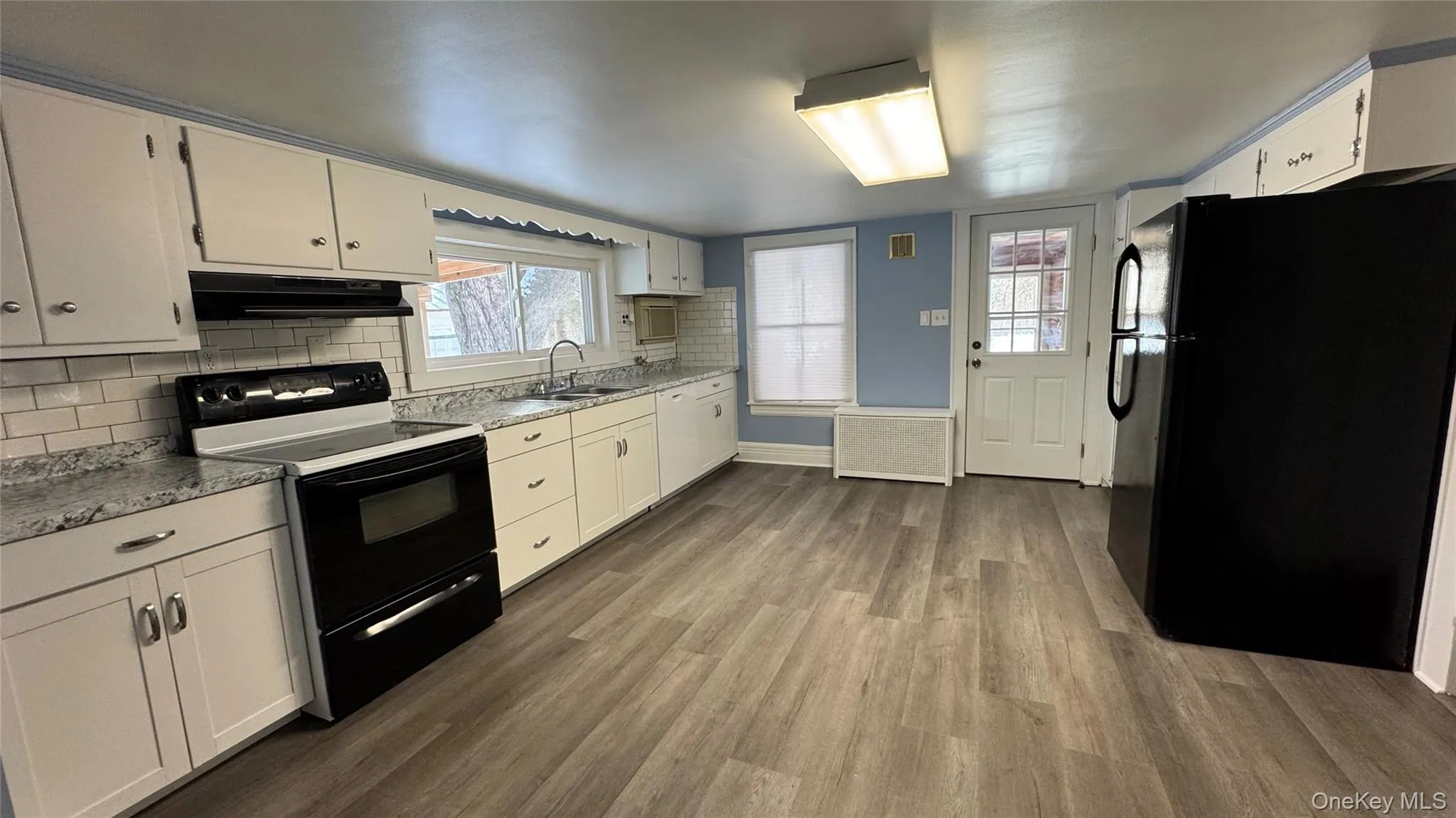 Kitchen featuring black appliances, dark wood-type flooring, radiator, white cabinetry, and under cabinet range hood Kitchen featuring black appliances, dark wood-type flooring, radiator, white cabinetry, and under cabinet range hood
