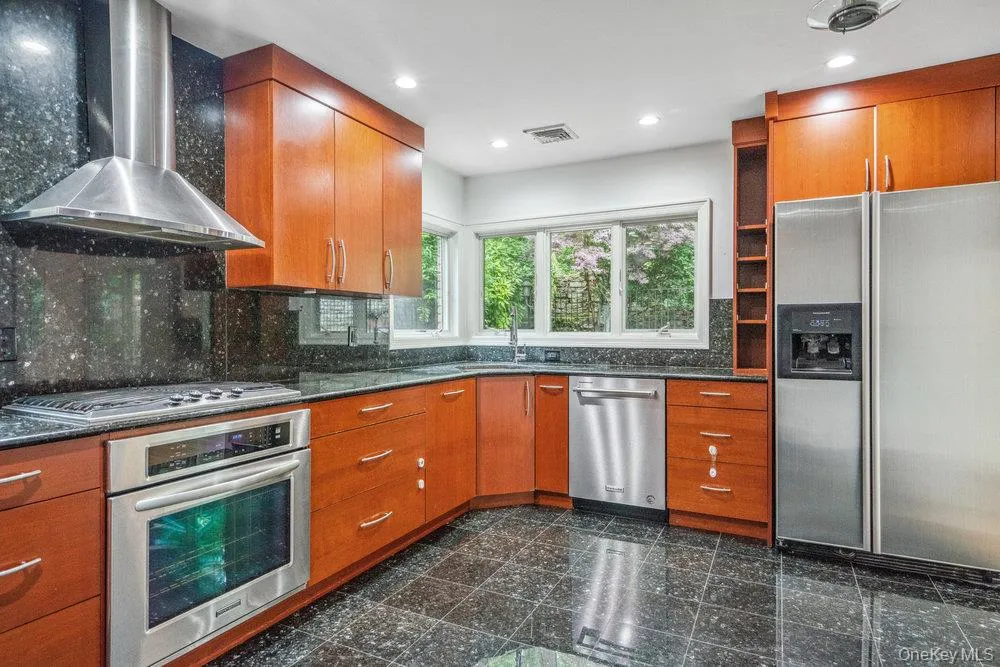 Kitchen featuring wall chimney exhaust hood, appliances with stainless steel finishes, granite tiled floors, brown cabinetry, and dark stone counters Kitchen featuring wall chimney exhaust hood, appliances with stainless steel finishes, granite tiled floors, brown cabinetry, and dark stone counters