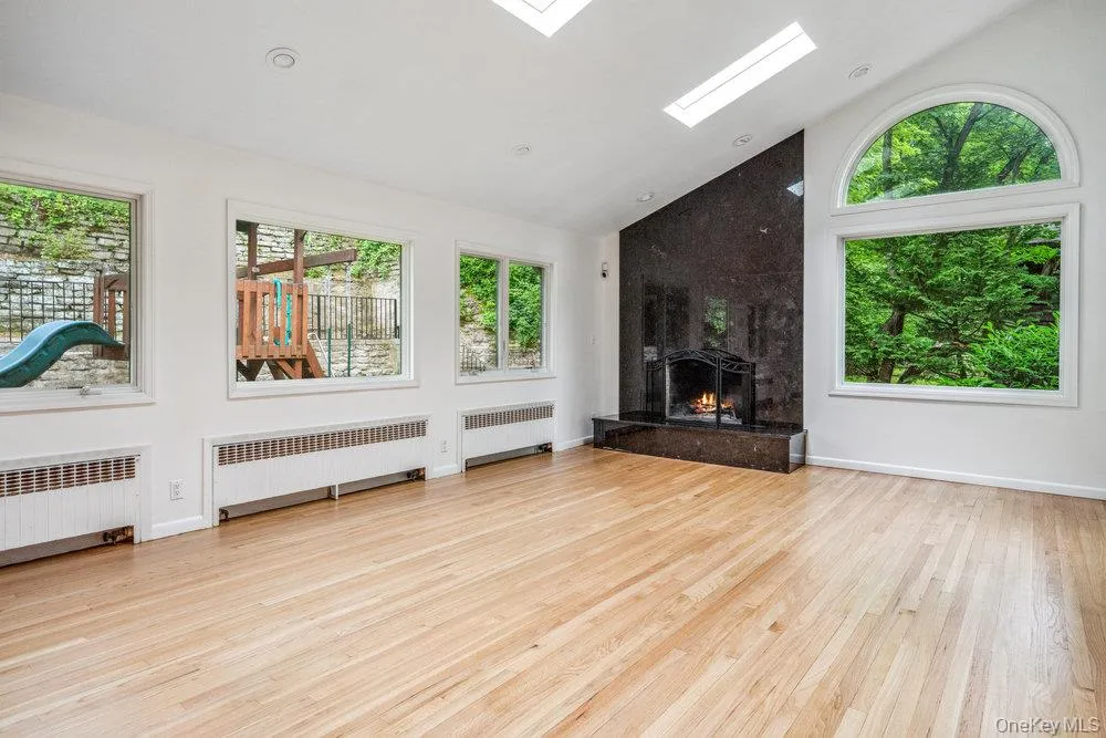 Unfurnished living room featuring radiator heating unit, a skylight, light wood-type flooring, a fireplace, and high vaulted ceiling Unfurnished living room featuring radiator heating unit, a skylight, light wood-type flooring, a fireplace, and high vaulted ceiling