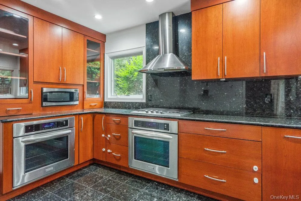 Kitchen featuring stainless steel appliances, range hood, granite floors, brown cabinetry, and dark stone countertops Kitchen featuring stainless steel appliances, range hood, granite floors, brown cabinetry, and dark stone countertops