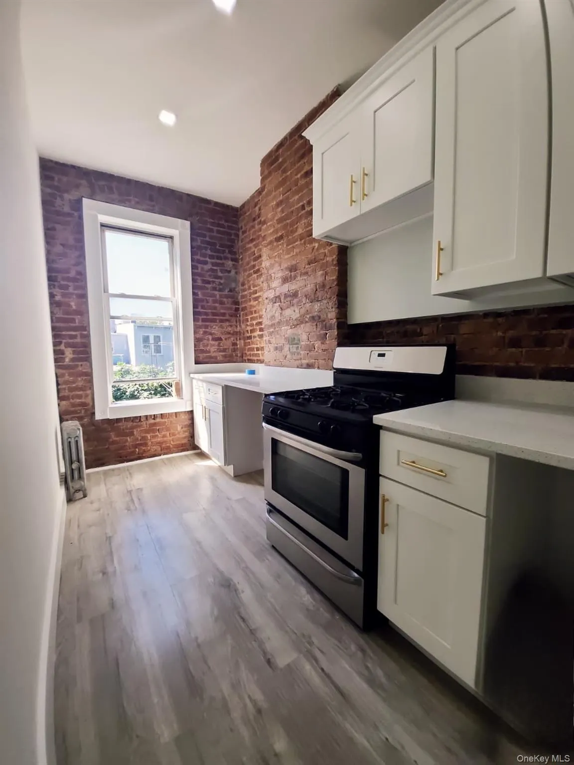 Kitchen with stainless steel gas range oven, white cabinets, brick wall, light wood finished floors, and light stone countertops Kitchen with stainless steel gas range oven, white cabinets, brick wall, light wood finished floors, and light stone countertops