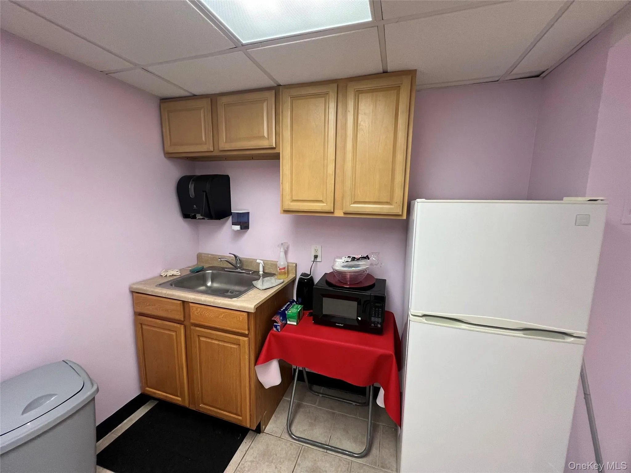 Kitchen featuring freestanding refrigerator, light countertops, a drop ceiling, light tile patterned flooring, and black microwave Kitchen featuring freestanding refrigerator, light countertops, a drop ceiling, light tile patterned flooring, and black microwave