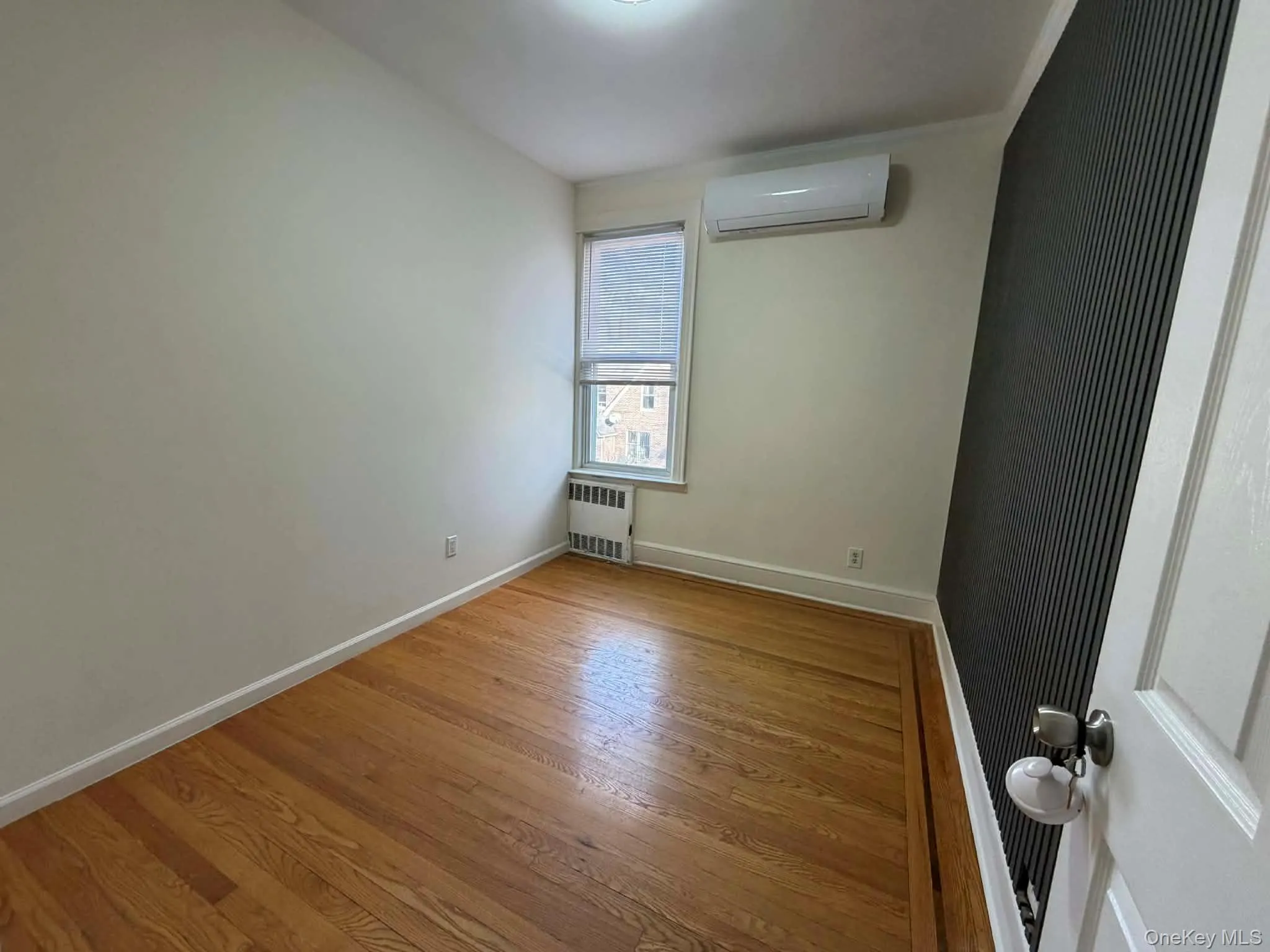Empty room featuring wood finished floors, a wall mounted air conditioner, and radiator heating unit Empty room featuring wood finished floors, a wall mounted air conditioner, and radiator heating unit