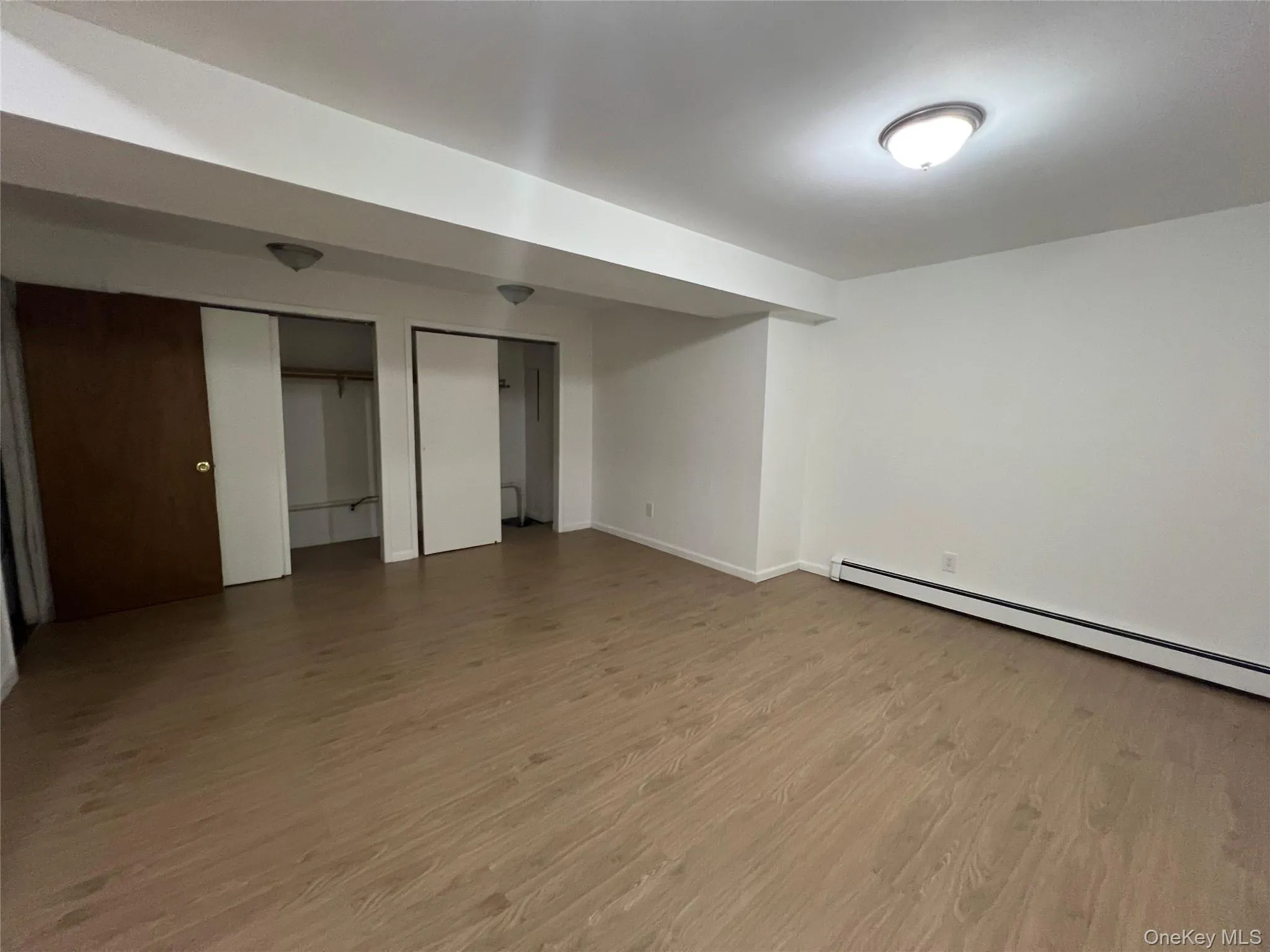 Unfurnished bedroom featuring multiple closets, light wood-type flooring, and a baseboard radiator Unfurnished bedroom featuring multiple closets, light wood-type flooring, and a baseboard radiator