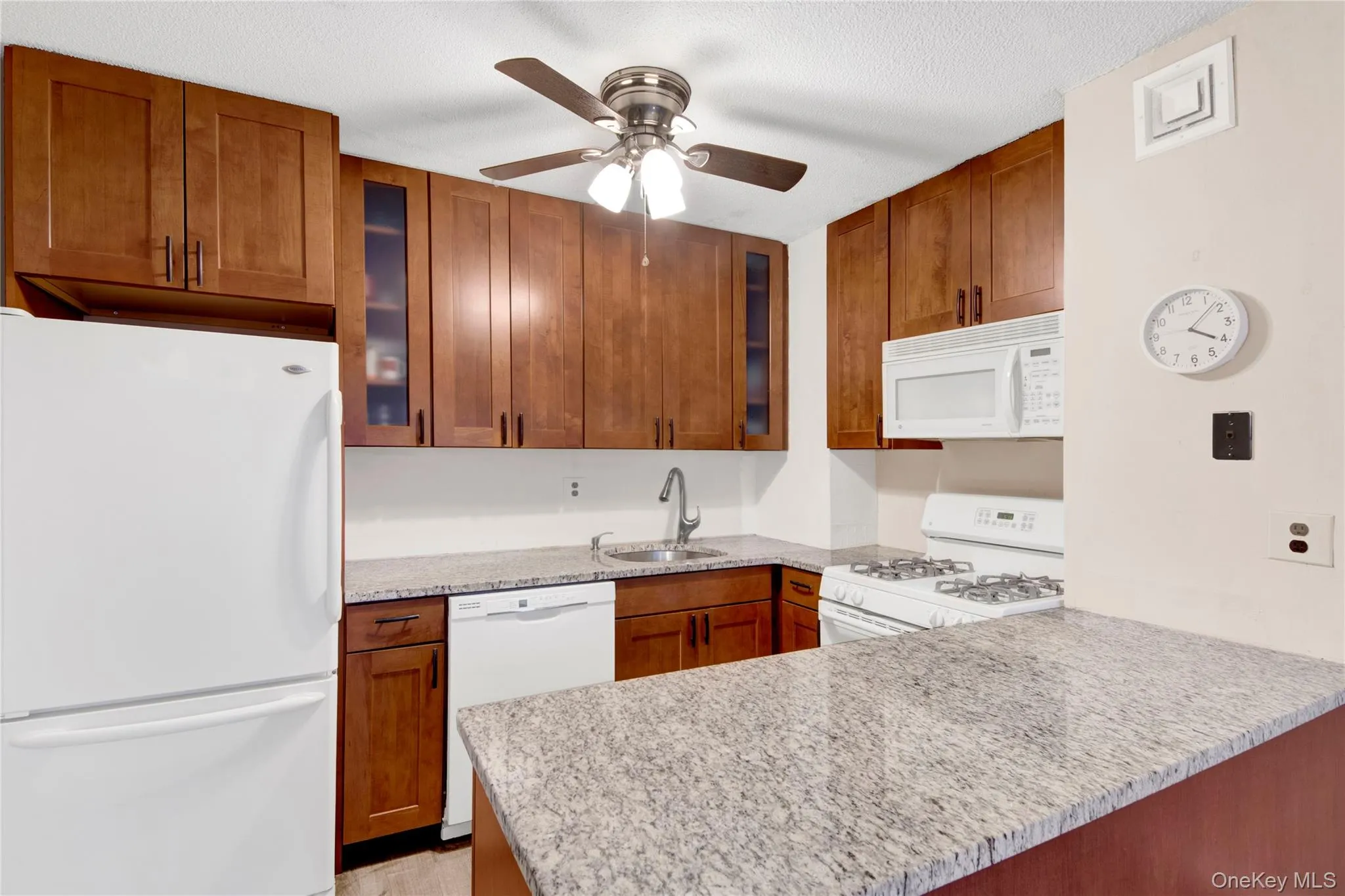 Kitchen featuring white appliances, brown cabinetry, glass insert cabinets, a ceiling fan, and light stone counters Kitchen featuring white appliances, brown cabinetry, glass insert cabinets, a ceiling fan, and light stone counters