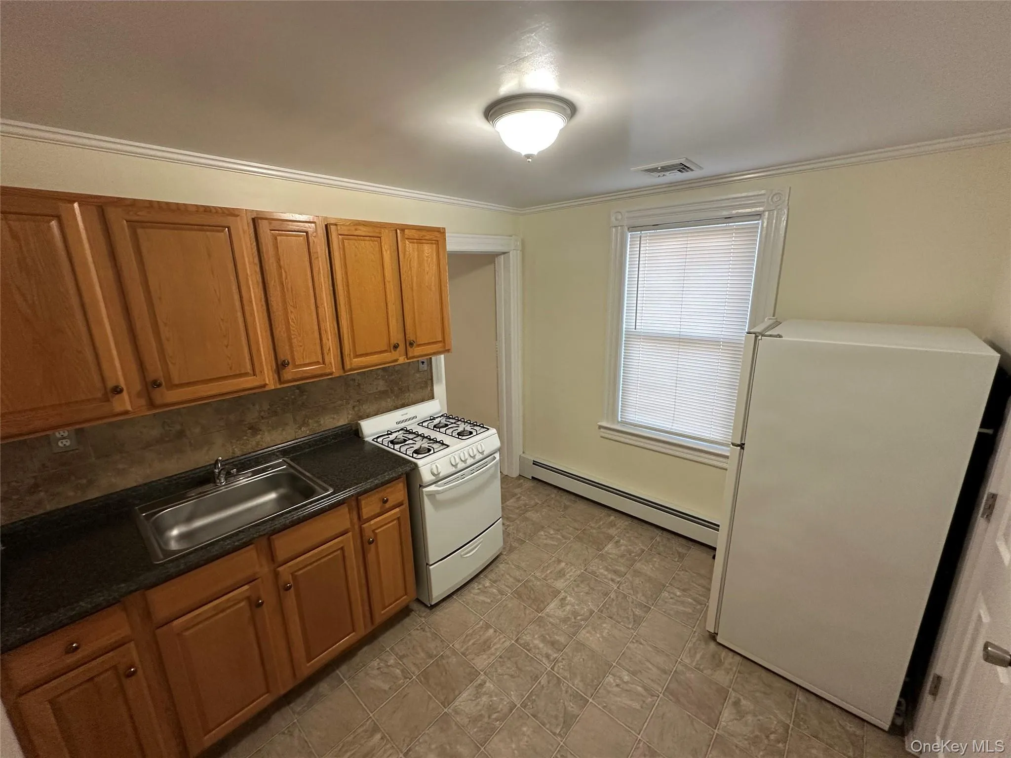 Kitchen featuring white appliances, dark countertops, brown cabinetry, baseboard heating, and ornamental molding Kitchen featuring white appliances, dark countertops, brown cabinetry, baseboard heating, and ornamental molding