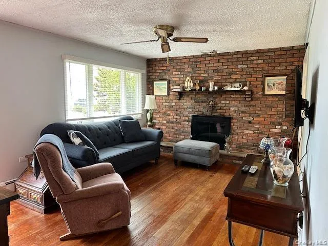 Living area featuring brick wall, wood-type flooring, a brick fireplace, a textured ceiling, and ceiling fan Living area featuring brick wall, wood-type flooring, a brick fireplace, a textured ceiling, and ceiling fan