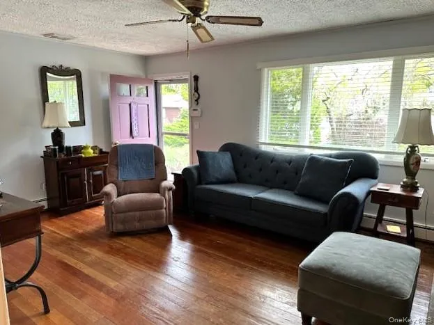 Living room featuring a textured ceiling, wood-type flooring, baseboard heating, ceiling fan, and healthy amount of natural light Living room featuring a textured ceiling, wood-type flooring, baseboard heating, ceiling fan, and healthy amount of natural light