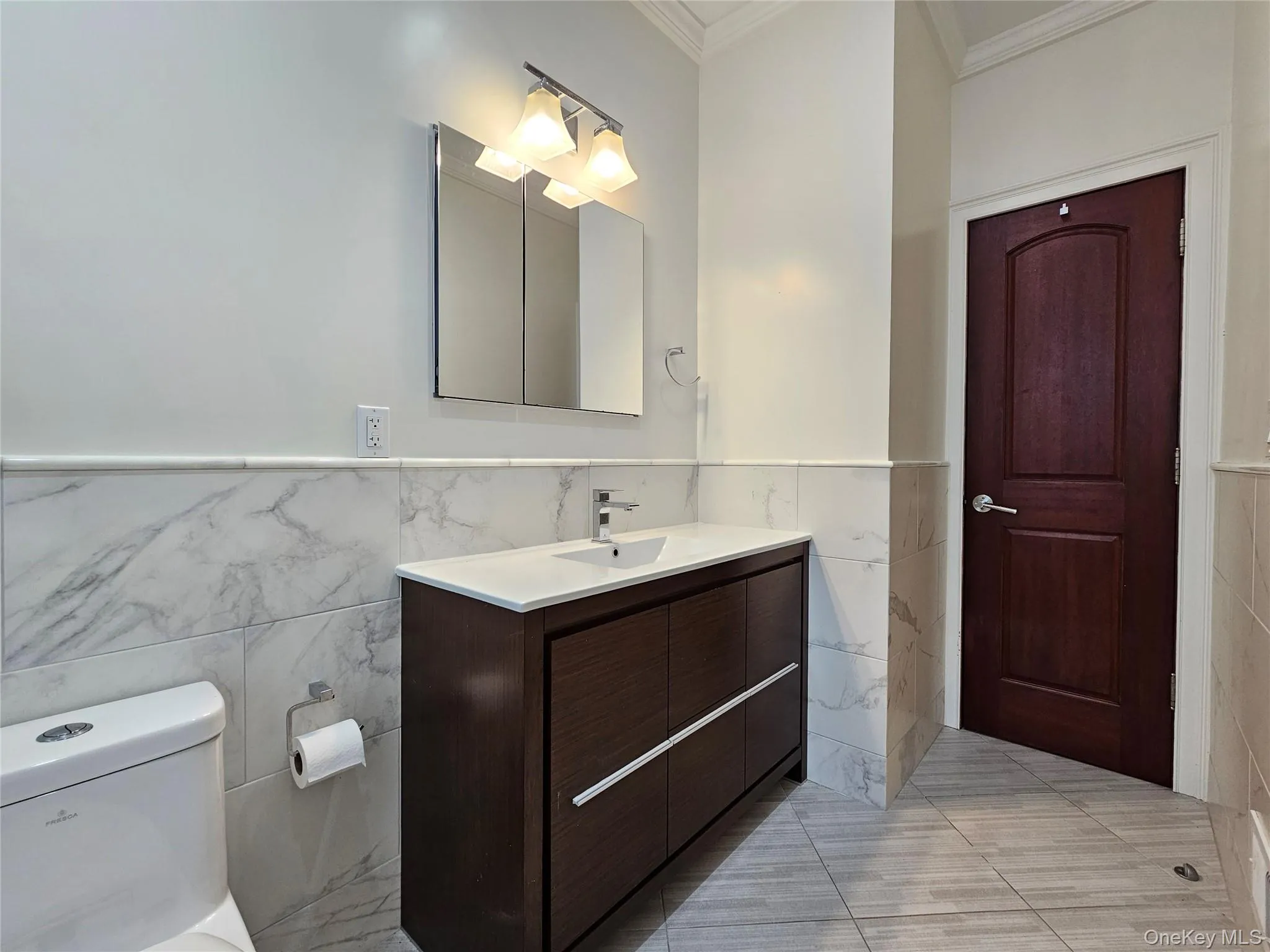 Bathroom featuring tile walls, a wainscoted wall, vanity, ornamental molding, and light tile patterned floors Bathroom featuring tile walls, a wainscoted wall, vanity, ornamental molding, and light tile patterned floors