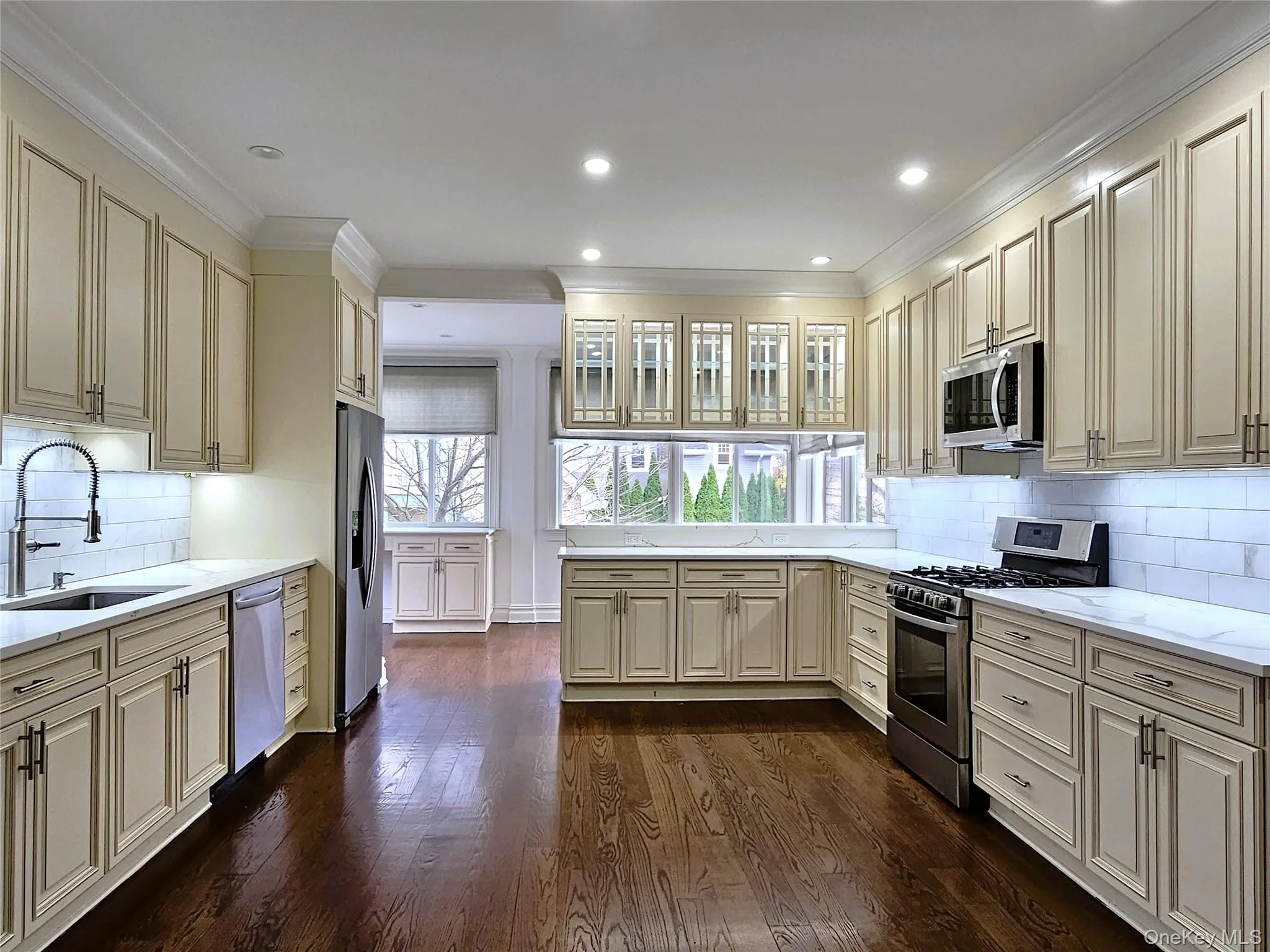 Kitchen featuring backsplash, cream cabinetry, appliances with stainless steel finishes, glass insert cabinets, and light stone counters Kitchen featuring backsplash, cream cabinetry, appliances with stainless steel finishes, glass insert cabinets, and light stone counters