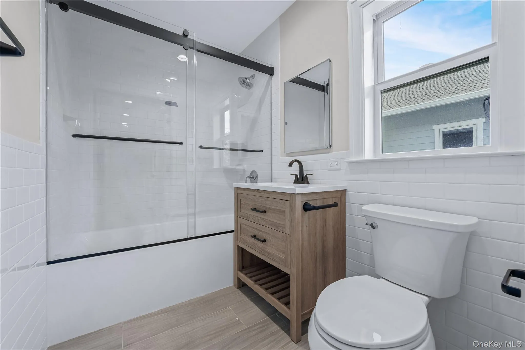 Bathroom featuring tile walls, bath / shower combo with glass door, a wainscoted wall, and vanity Bathroom featuring tile walls, bath / shower combo with glass door, a wainscoted wall, and vanity