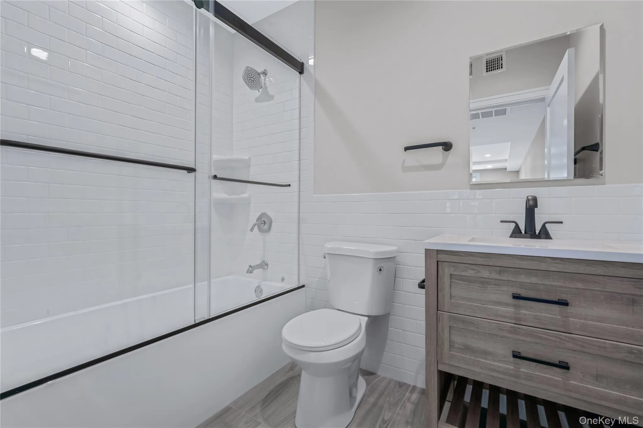Bathroom with tile walls, vanity, bath / shower combo with glass door, and a wainscoted wall Bathroom with tile walls, vanity, bath / shower combo with glass door, and a wainscoted wall