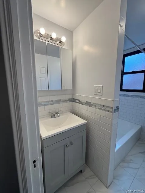 Full bathroom with vanity, light marble finish floors, wainscoting, tile walls, and shower / bath combination Full bathroom with vanity, light marble finish floors, wainscoting, tile walls, and shower / bath combination