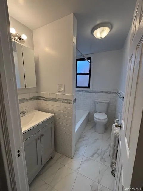 Full bathroom featuring vanity, wainscoting, tile walls, light marble finish flooring, and radiator Full bathroom featuring vanity, wainscoting, tile walls, light marble finish flooring, and radiator