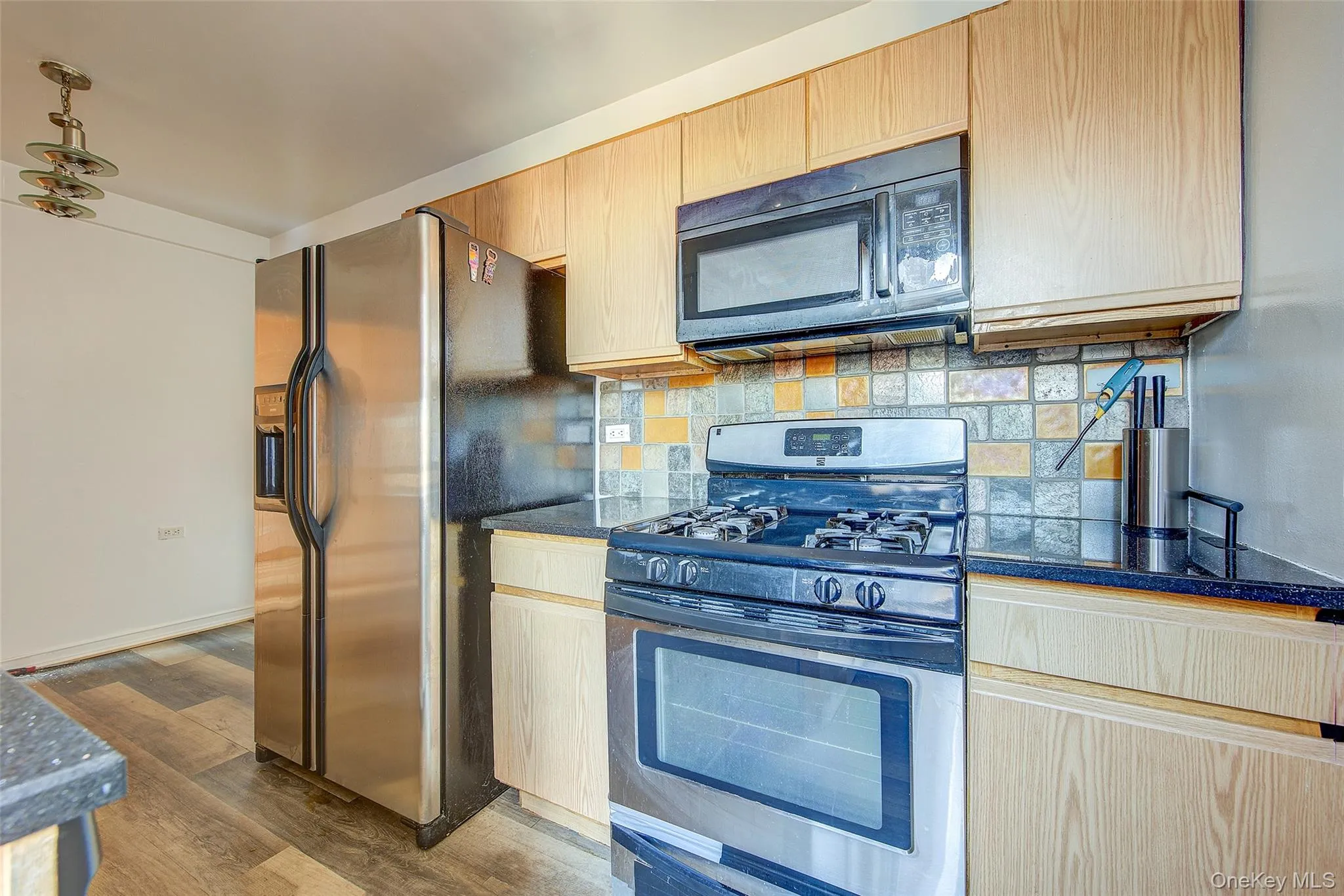 Kitchen with stainless steel appliances, light brown cabinets, tasteful backsplash, dark wood-style flooring, and dark stone countertops Kitchen with stainless steel appliances, light brown cabinets, tasteful backsplash, dark wood-style flooring, and dark stone countertops