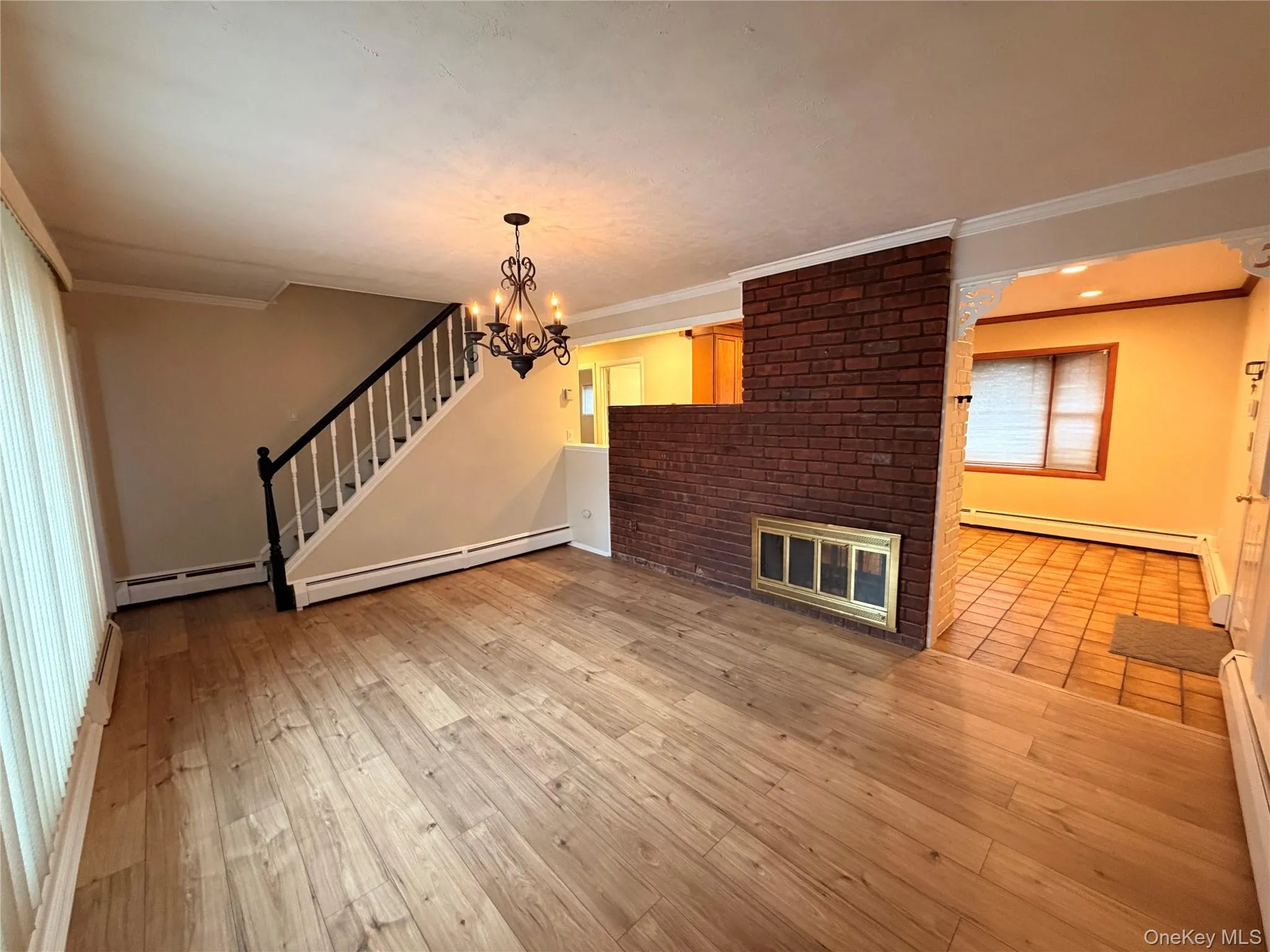 Unfurnished living room with crown molding, a brick fireplace, baseboard heating, and stairs Unfurnished living room with crown molding, a brick fireplace, baseboard heating, and stairs