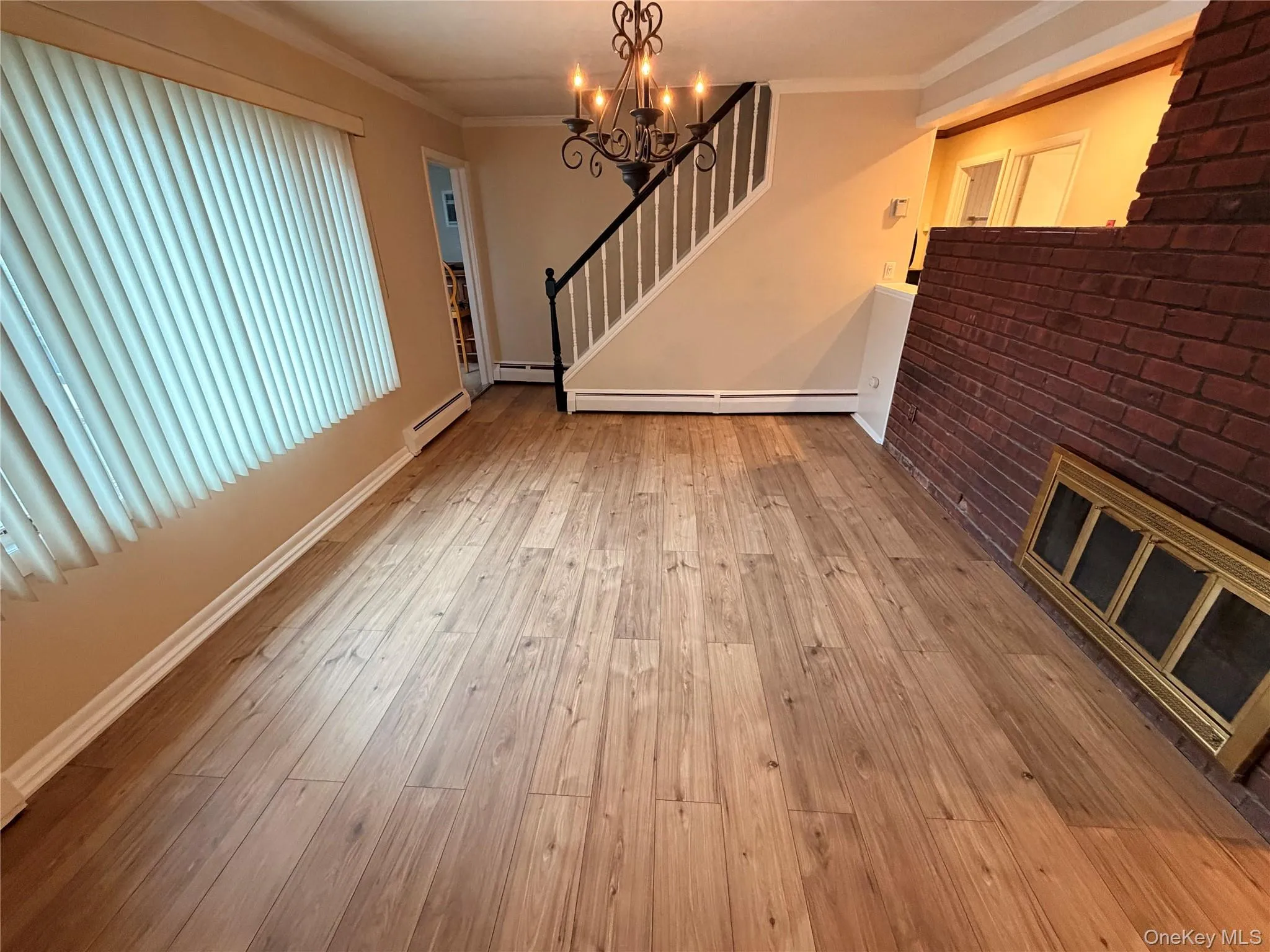 Unfurnished living room featuring a chandelier, ornamental molding, light wood finished floors, a baseboard radiator, and stairs Unfurnished living room featuring a chandelier, ornamental molding, light wood finished floors, a baseboard radiator, and stairs