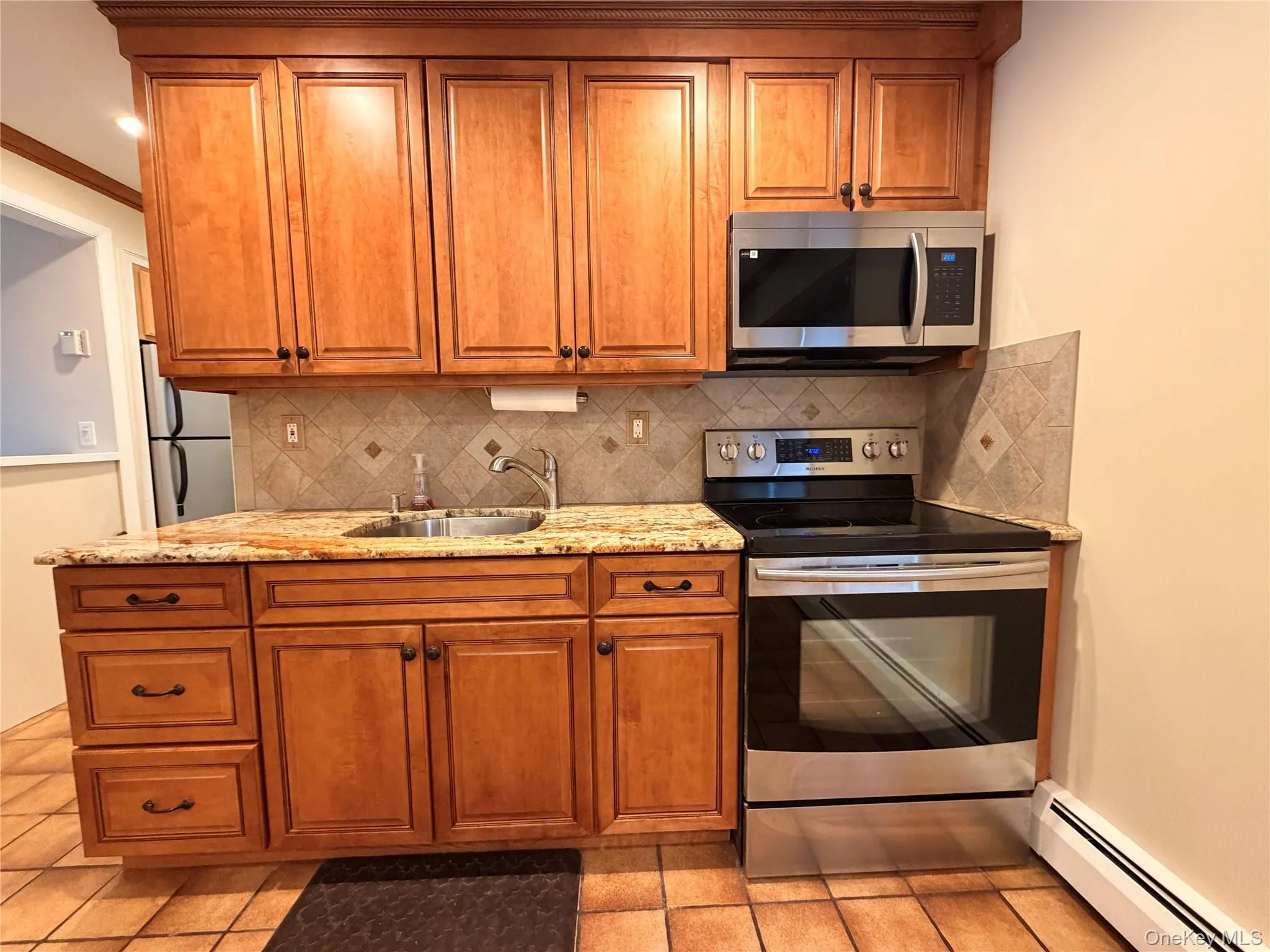 Kitchen featuring appliances with stainless steel finishes, brown cabinets, a baseboard radiator, light stone countertops, and tasteful backsplash Kitchen featuring appliances with stainless steel finishes, brown cabinets, a baseboard radiator, light stone countertops, and tasteful backsplash