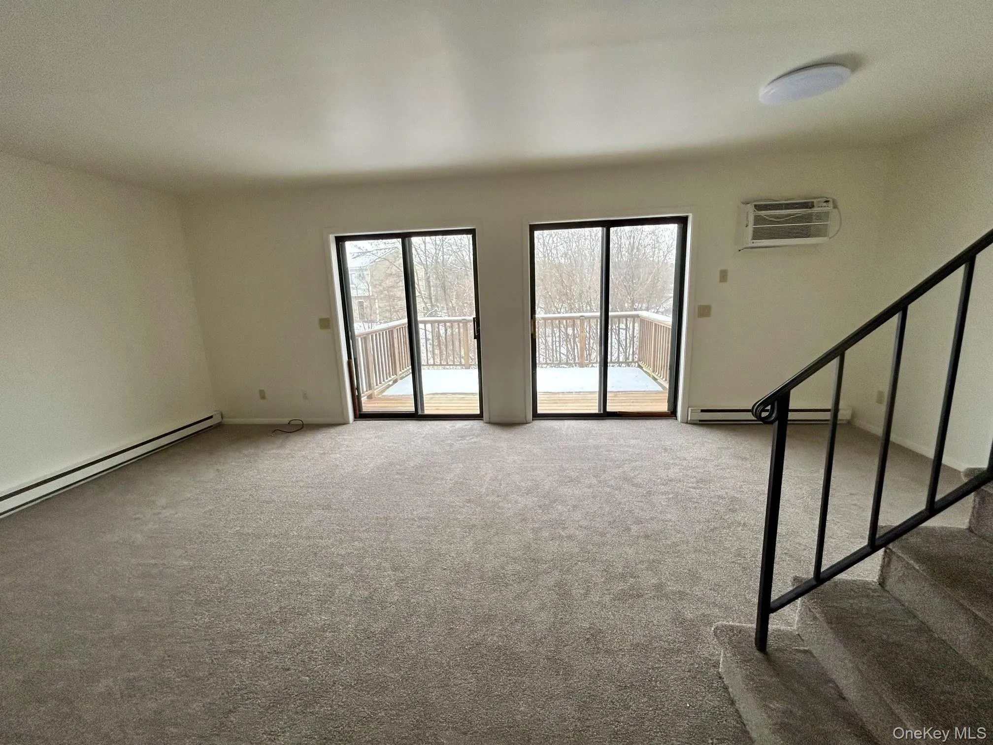 Unfurnished living room featuring light colored carpet, stairway, an AC wall unit, and baseboard heating Unfurnished living room featuring light colored carpet, stairway, an AC wall unit, and baseboard heating