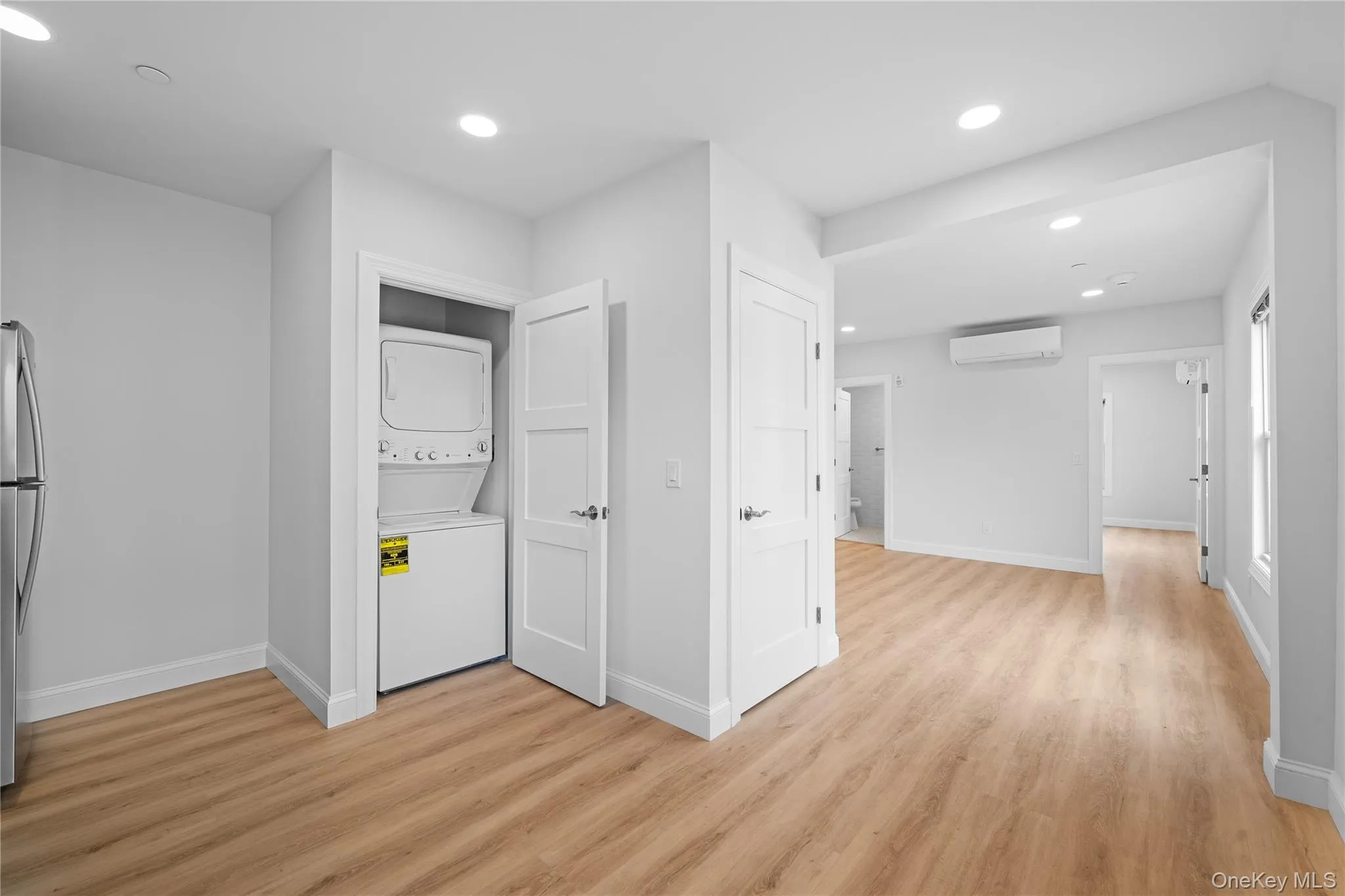 Washroom featuring stacked washer / dryer, recessed lighting, light wood-style flooring, and an AC wall unit Washroom featuring stacked washer / dryer, recessed lighting, light wood-style flooring, and an AC wall unit