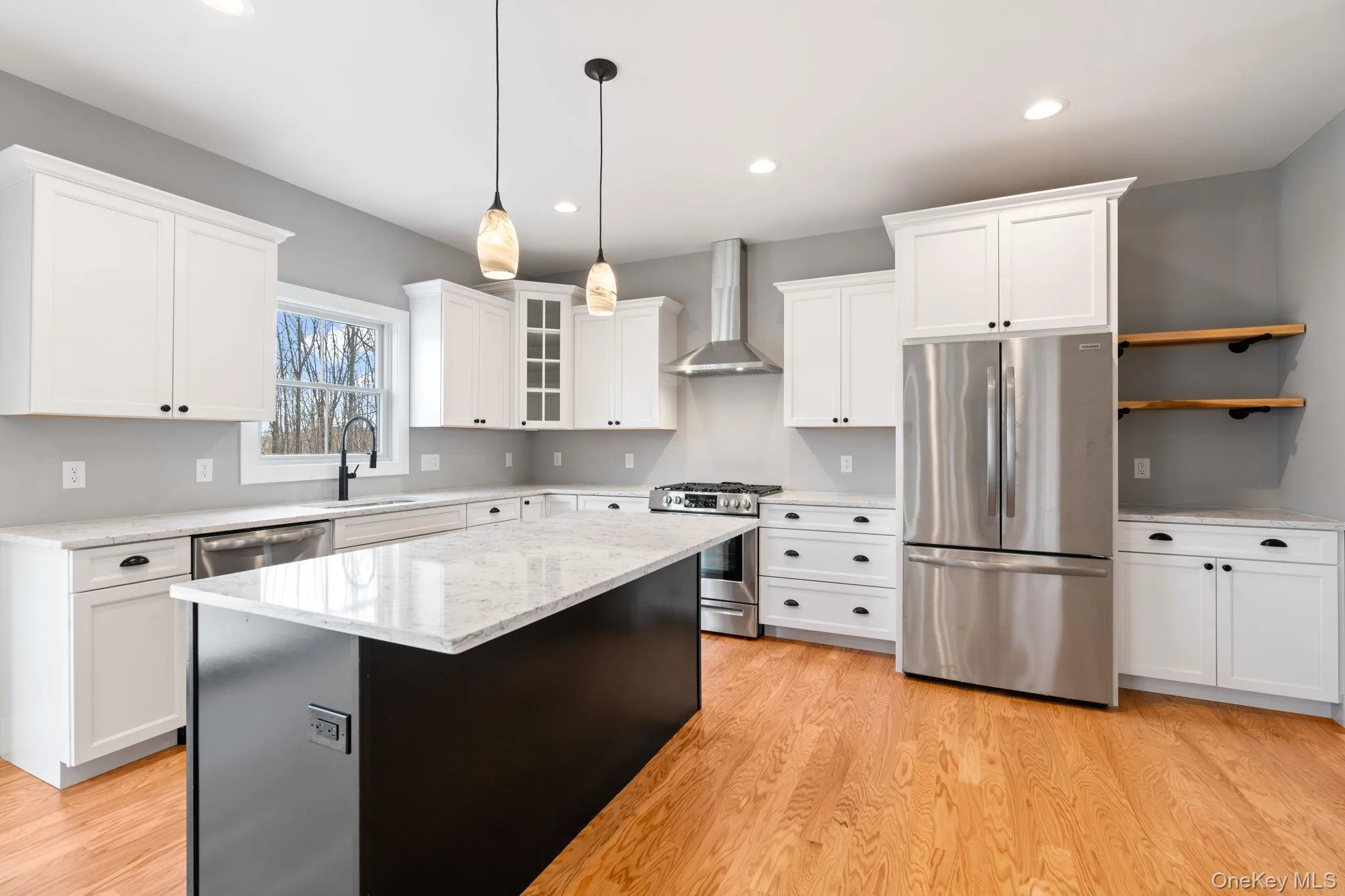 Kitchen with stainless steel appliances, glass insert cabinets, wall chimney range hood, white cabinetry, and hanging light fixtures Kitchen with stainless steel appliances, glass insert cabinets, wall chimney range hood, white cabinetry, and hanging light fixtures