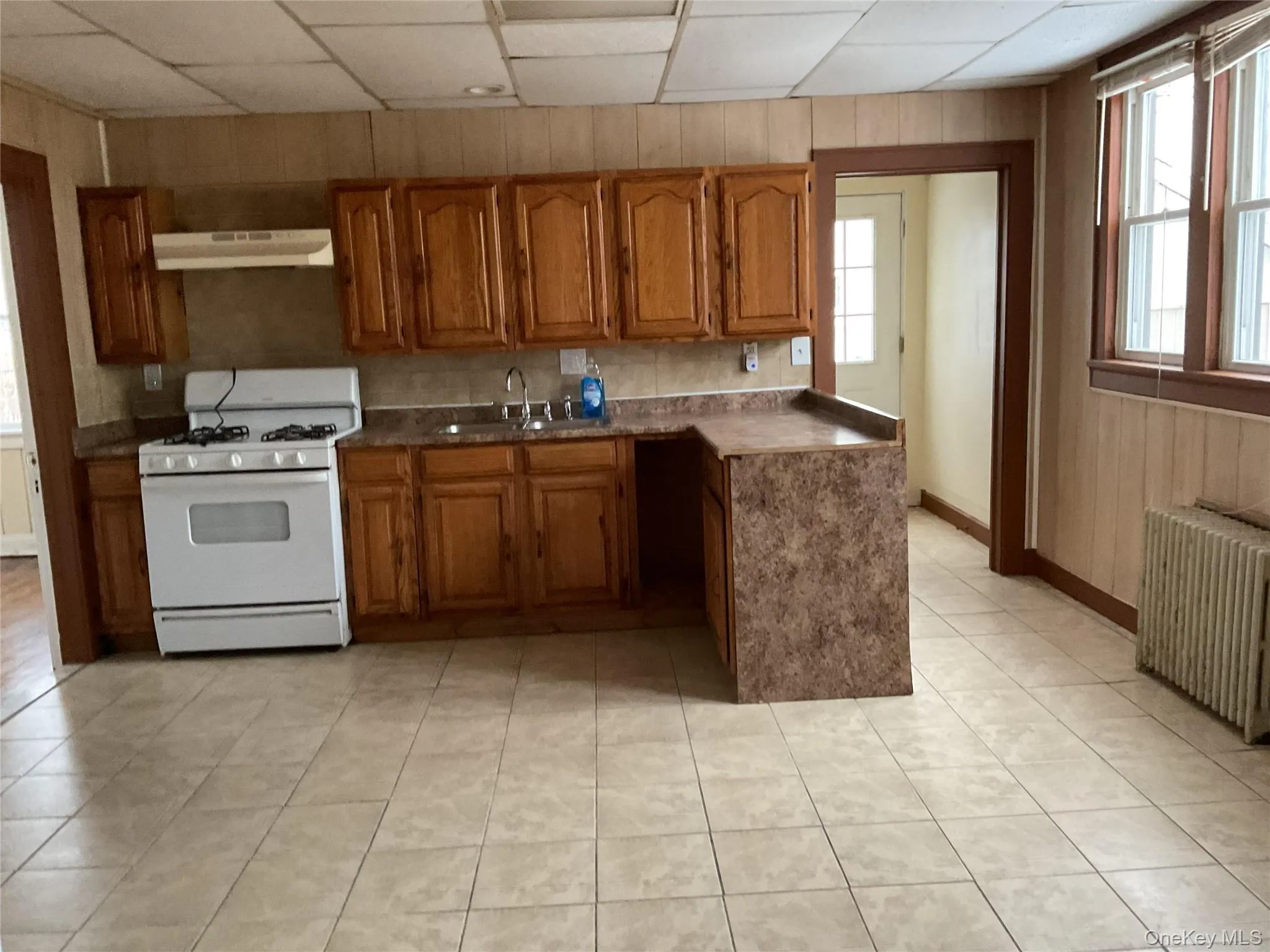 Kitchen with brown cabinetry, gas range gas stove, wooden walls, radiator, and ventilation hood Kitchen with brown cabinetry, gas range gas stove, wooden walls, radiator, and ventilation hood