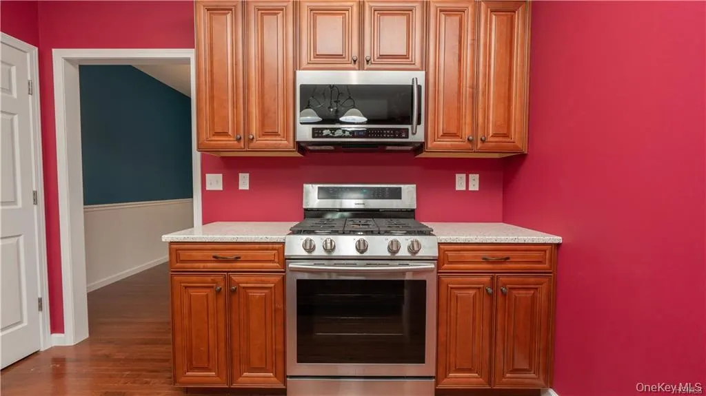 Kitchen featuring stainless steel appliances, brown cabinetry, dark wood finished floors, and light stone counters Kitchen featuring stainless steel appliances, brown cabinetry, dark wood finished floors, and light stone counters